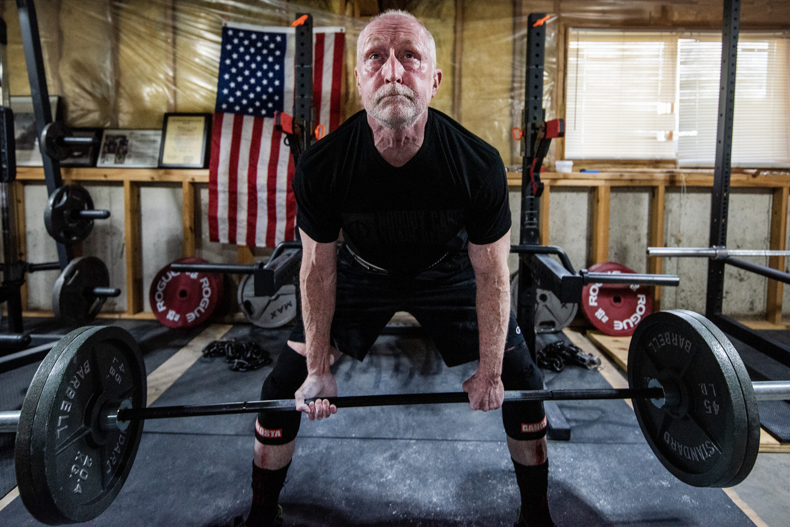Michael Love's face is red as he pulls up on a bar with two large weights on either side. He's in his basement with tan walls and a U.S. flag behind him.