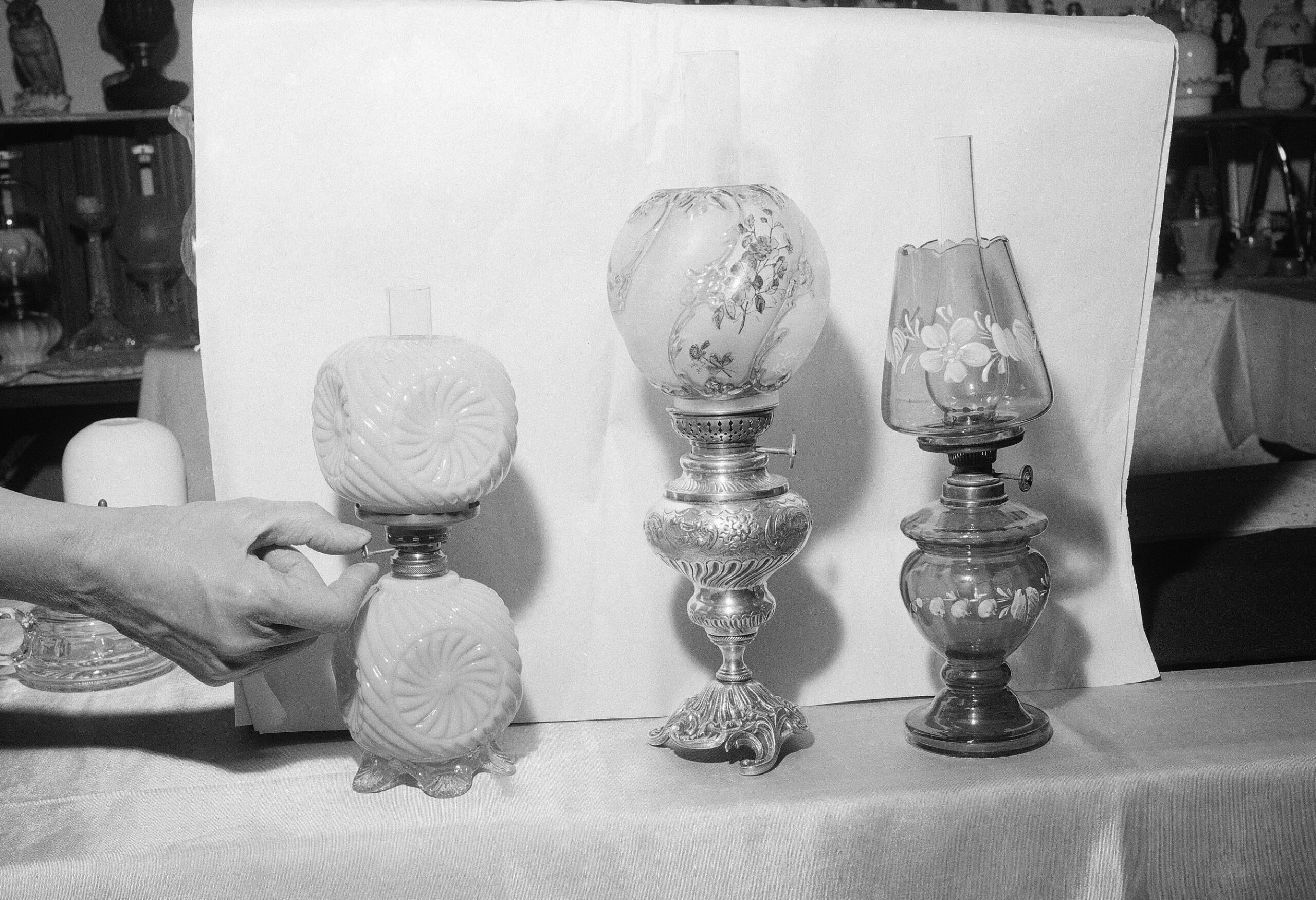 Three lamps sit in a cloth-covered table. A hand reaches out to touch one. The photo is black and white.