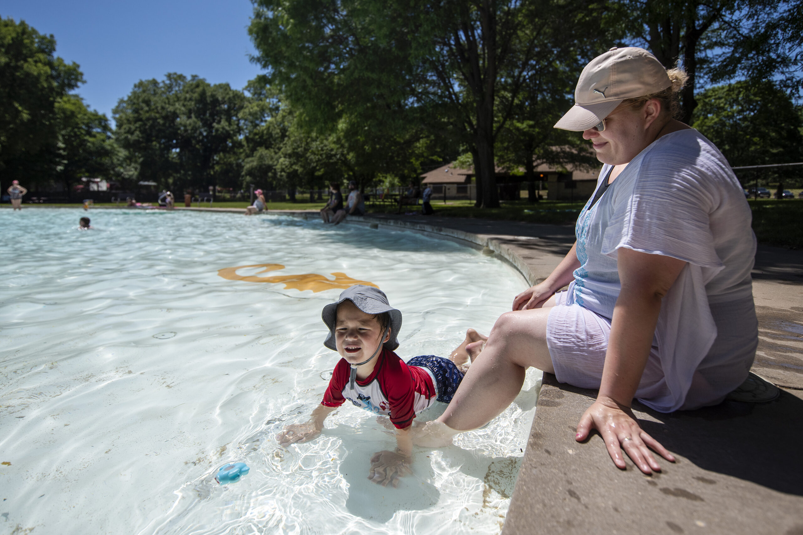 A child in a swimsuit and hat crawls around a shallow splash pool while his mother sits on the ledge nearby.