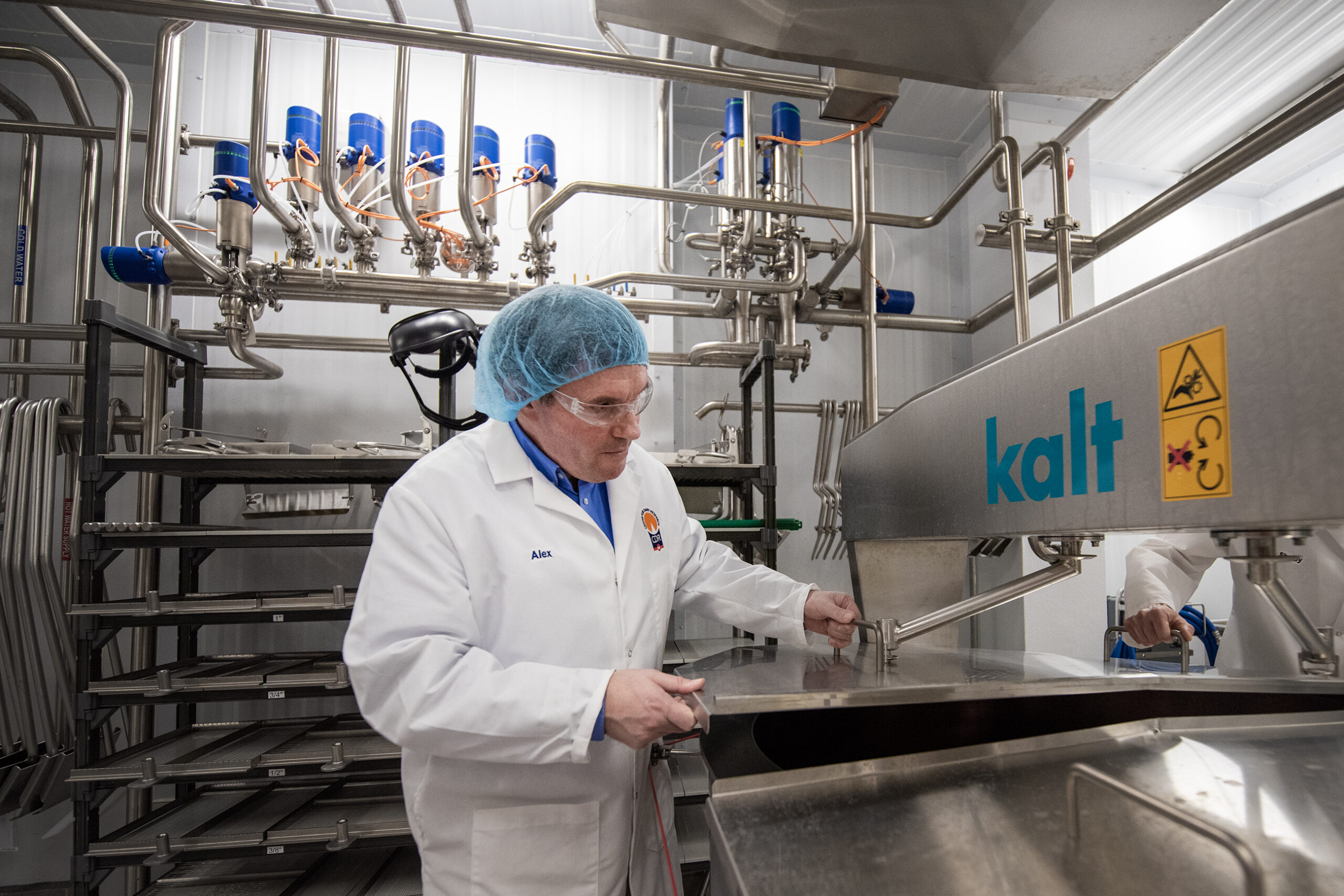 A man in a white lab coat lifts a lid on a piece of stainless steel machinery.