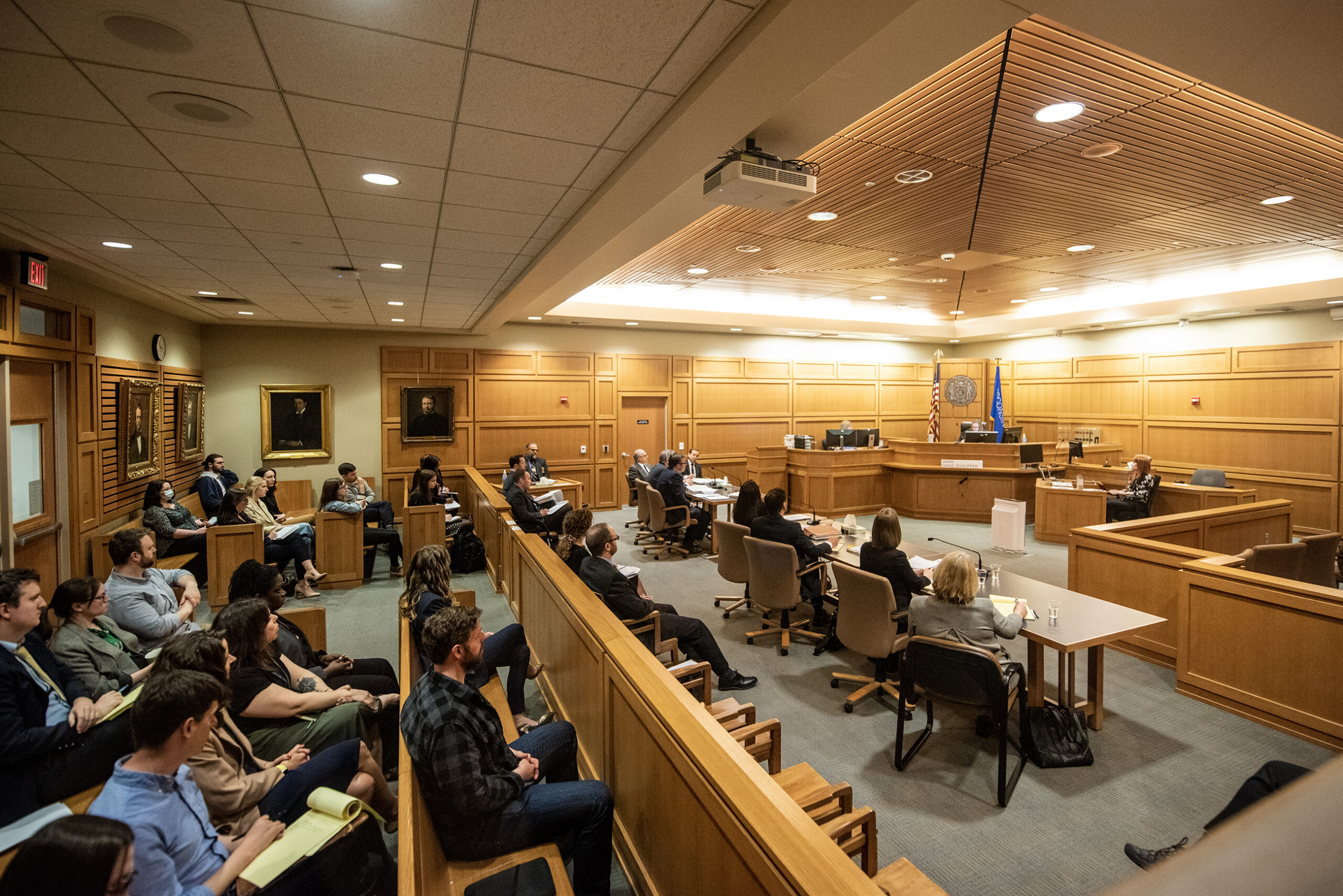 Several attorneys, reporters, and other people sit in the courtroom.