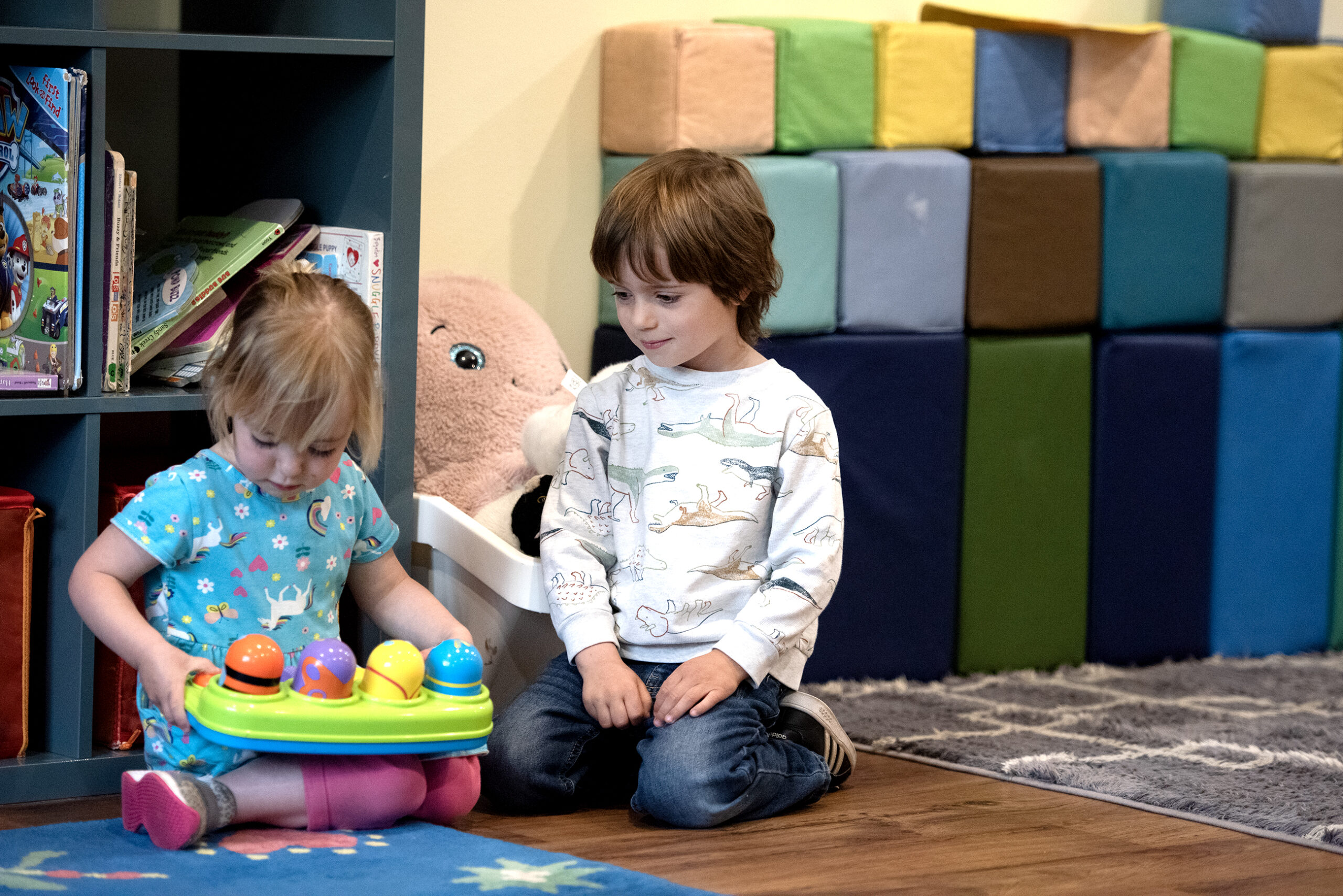 Two children sit on the floor of a classroom as they play with toys.