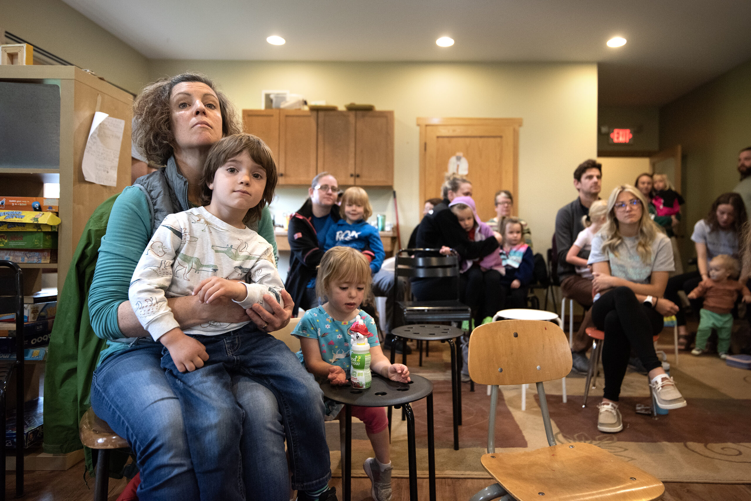 A mother holds a child in her lap while in a classroom with other parents, children and workers.