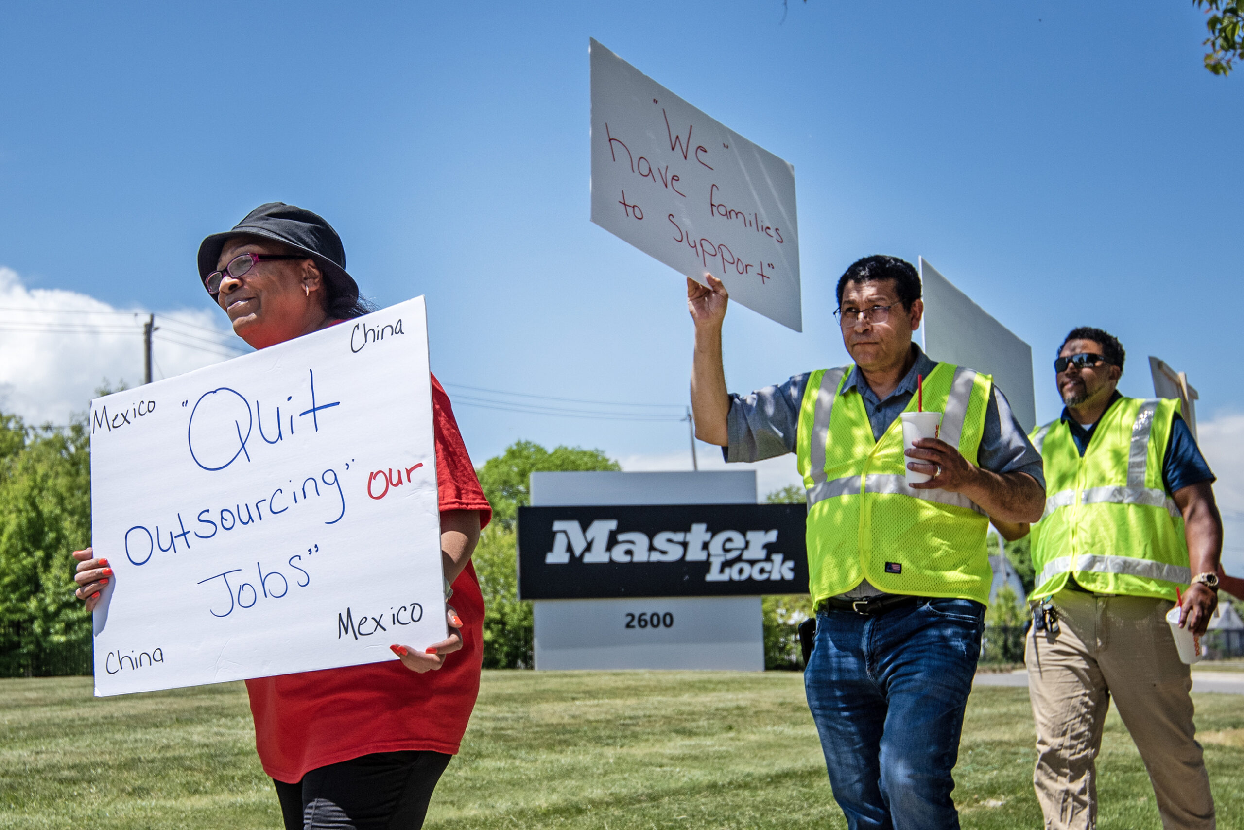 Three people walk in front of the large Master Lock sign outside of the plant. A woman's sign reads "Quit outsourcing our jobs."