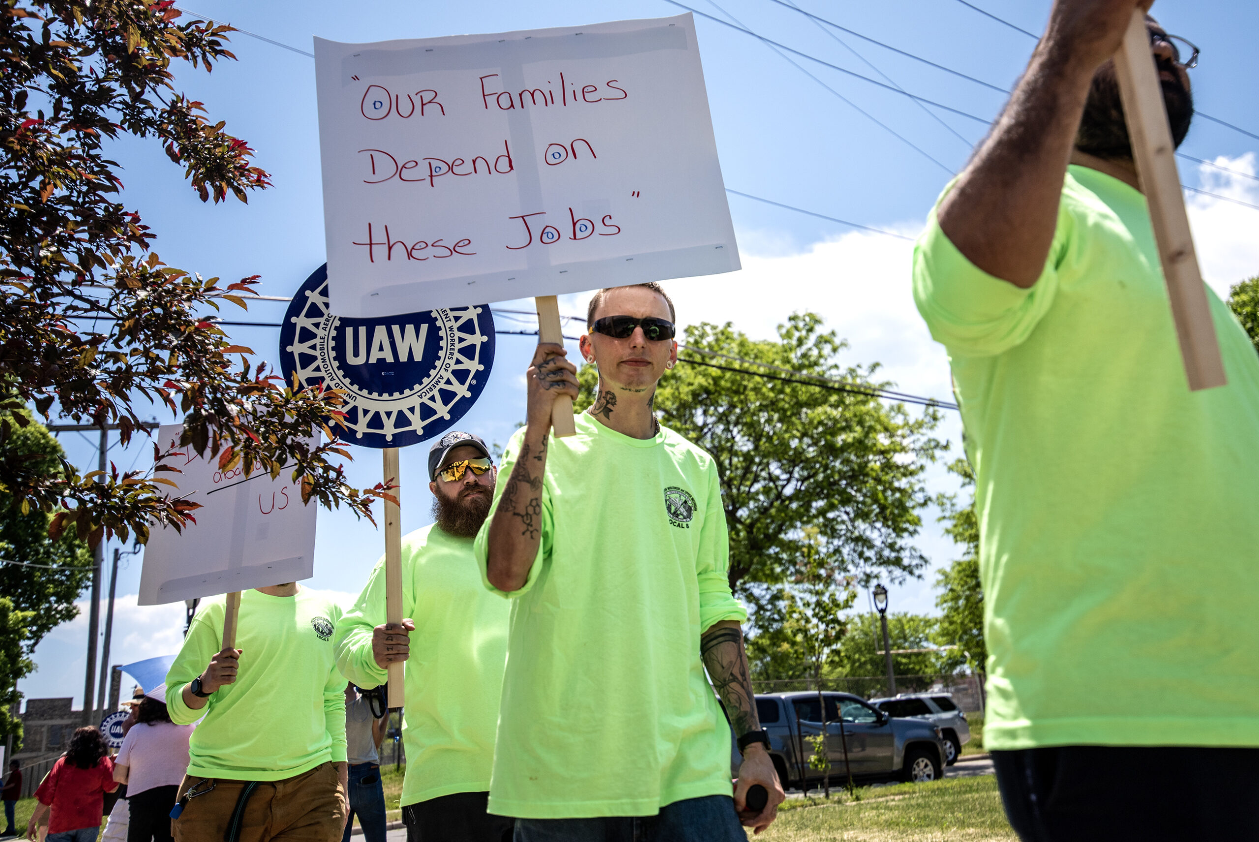 Workers in neon yellow shirts walk in a line. A sign held by one of the protesters says, 