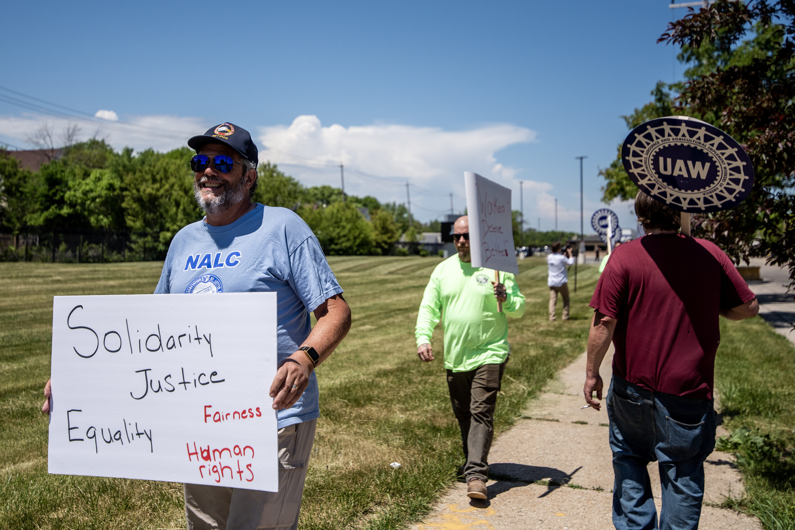 Several people can be seen on a sidewalk on a sunny day. A sign held by a protester says 