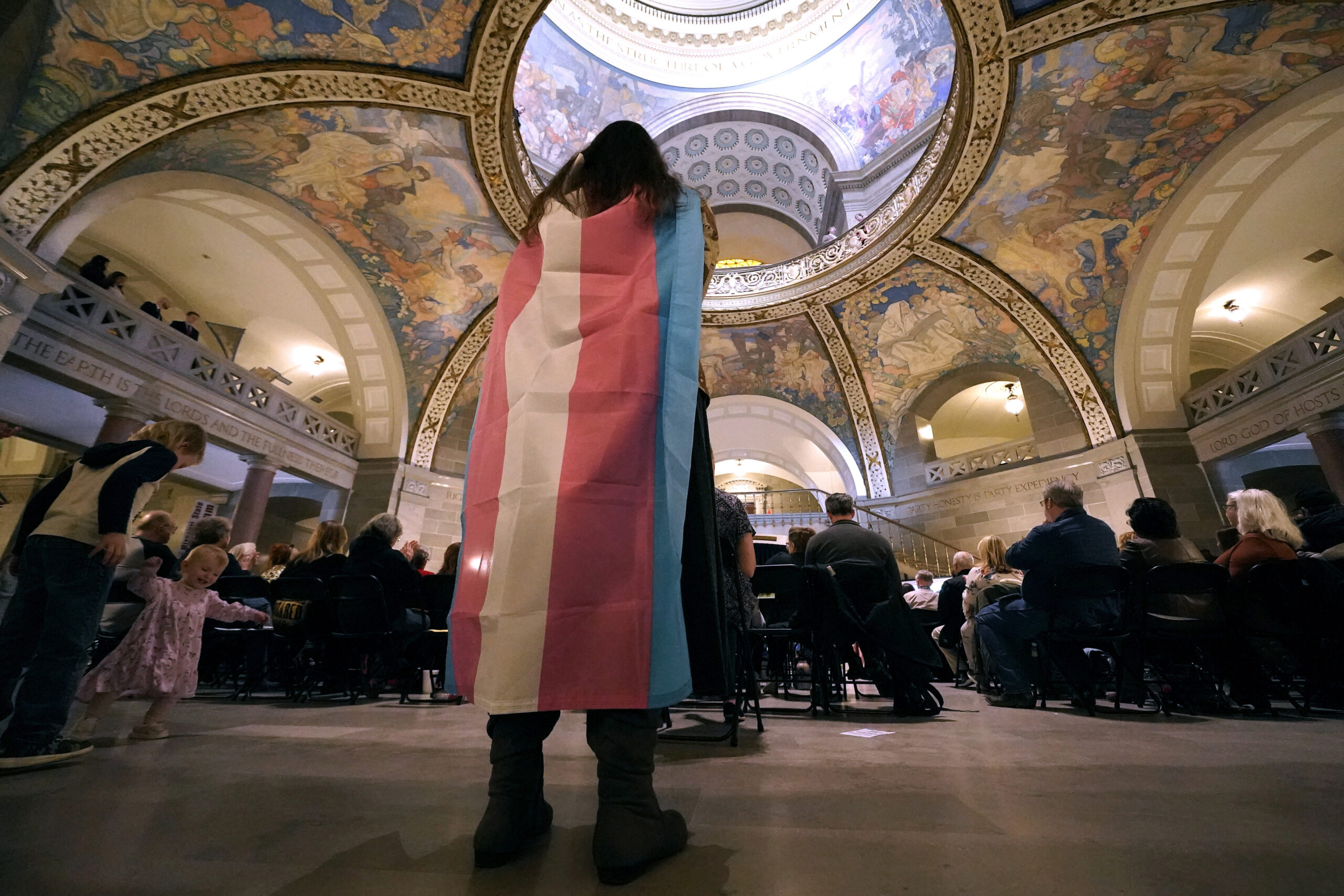 Someone with a transgender flag draped on their back stands in the Missouri statehouse