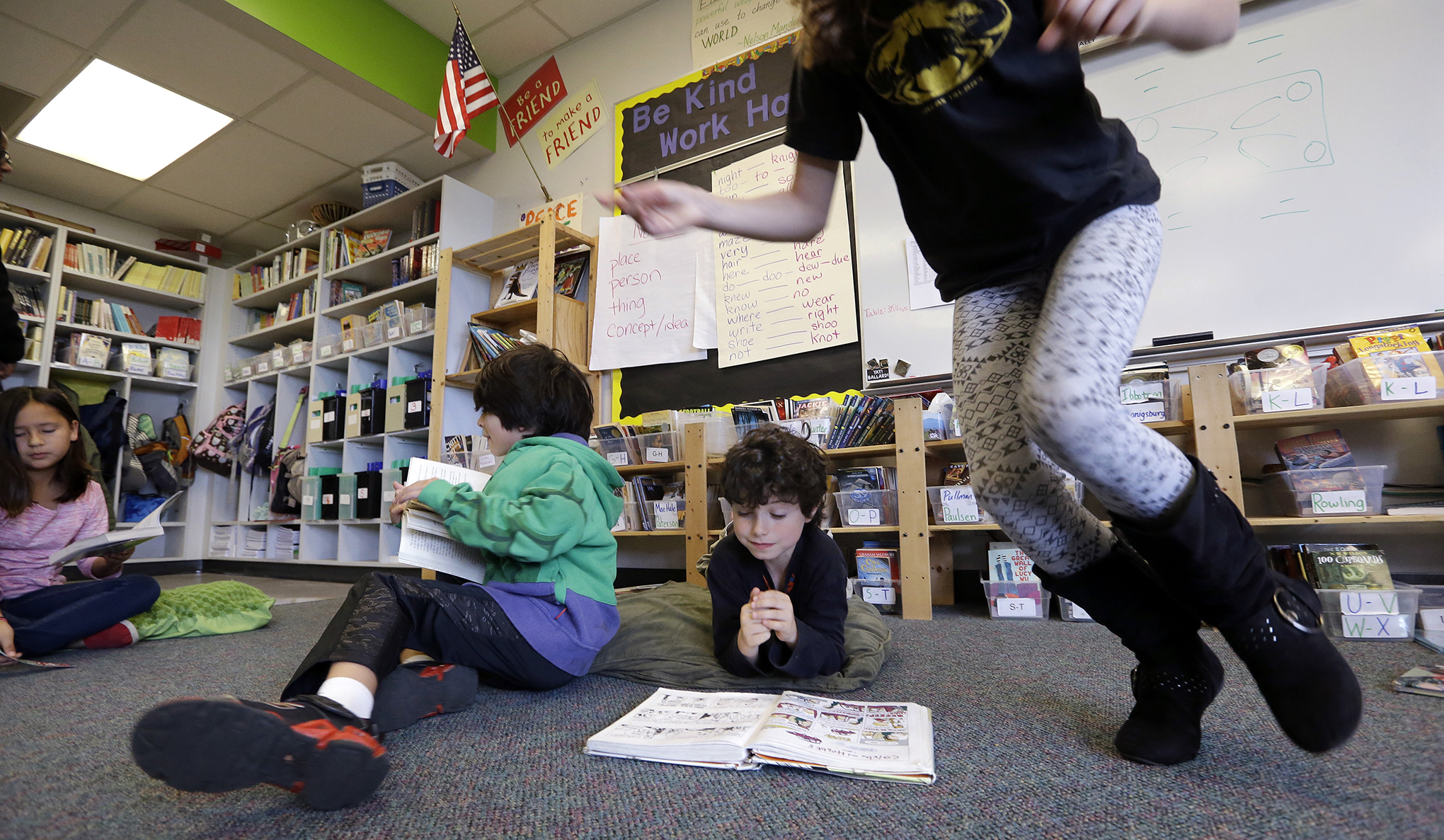 A fourth grade girl jumps up and runs past a boy who is reading.