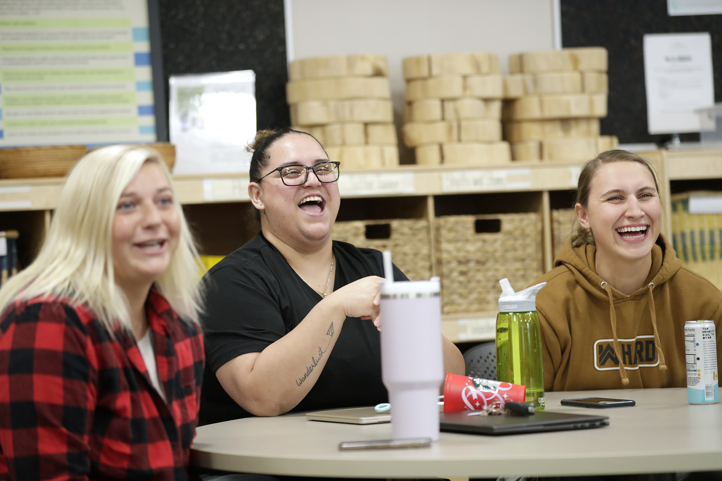 Three people sit at a table in a classroom, laughing