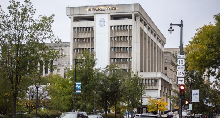 Cars and pedestrians pass by in front of a building with "Milwaukee Police" written on the outside.