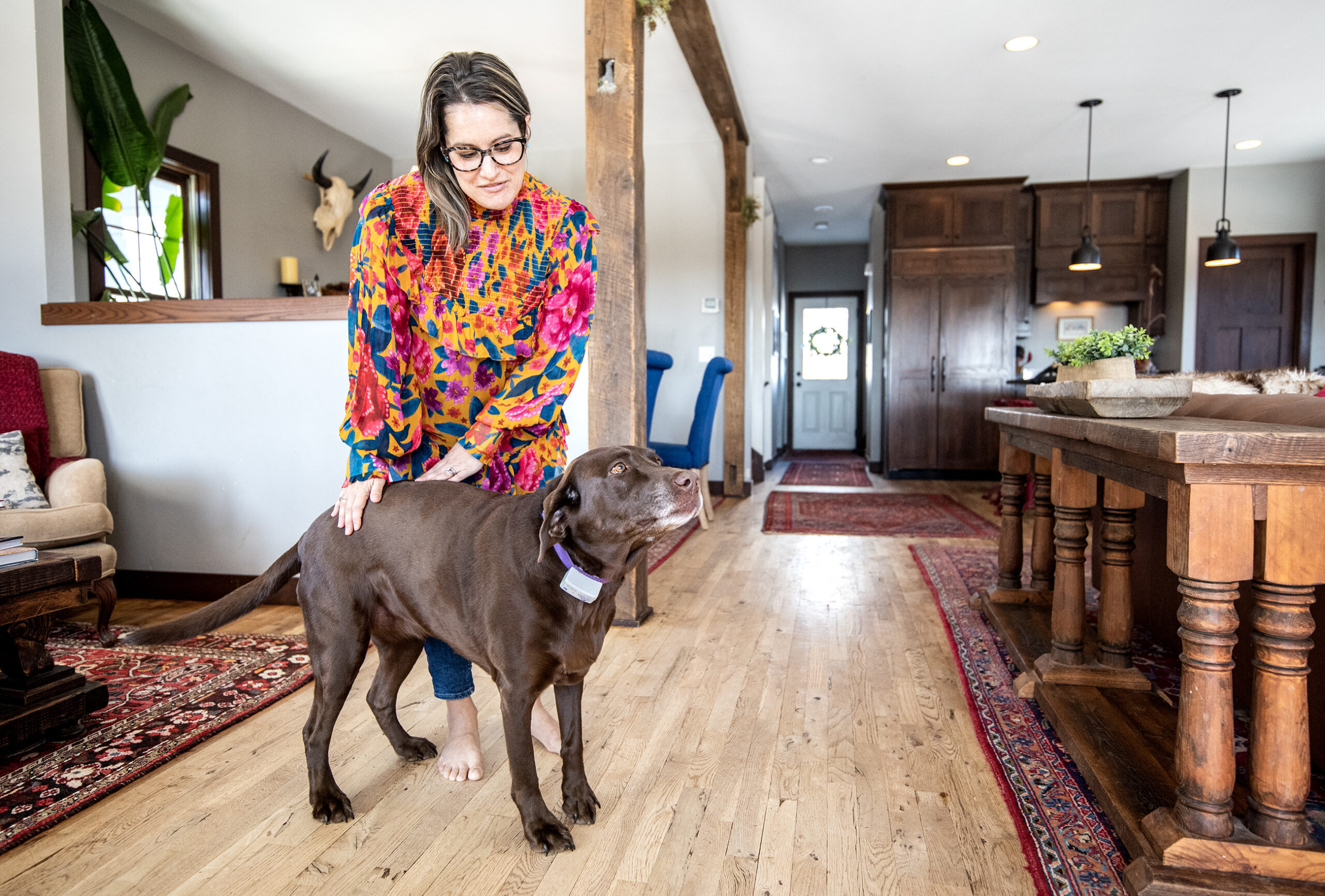 A woman stands in her home and leans down to pet her brown dog.
