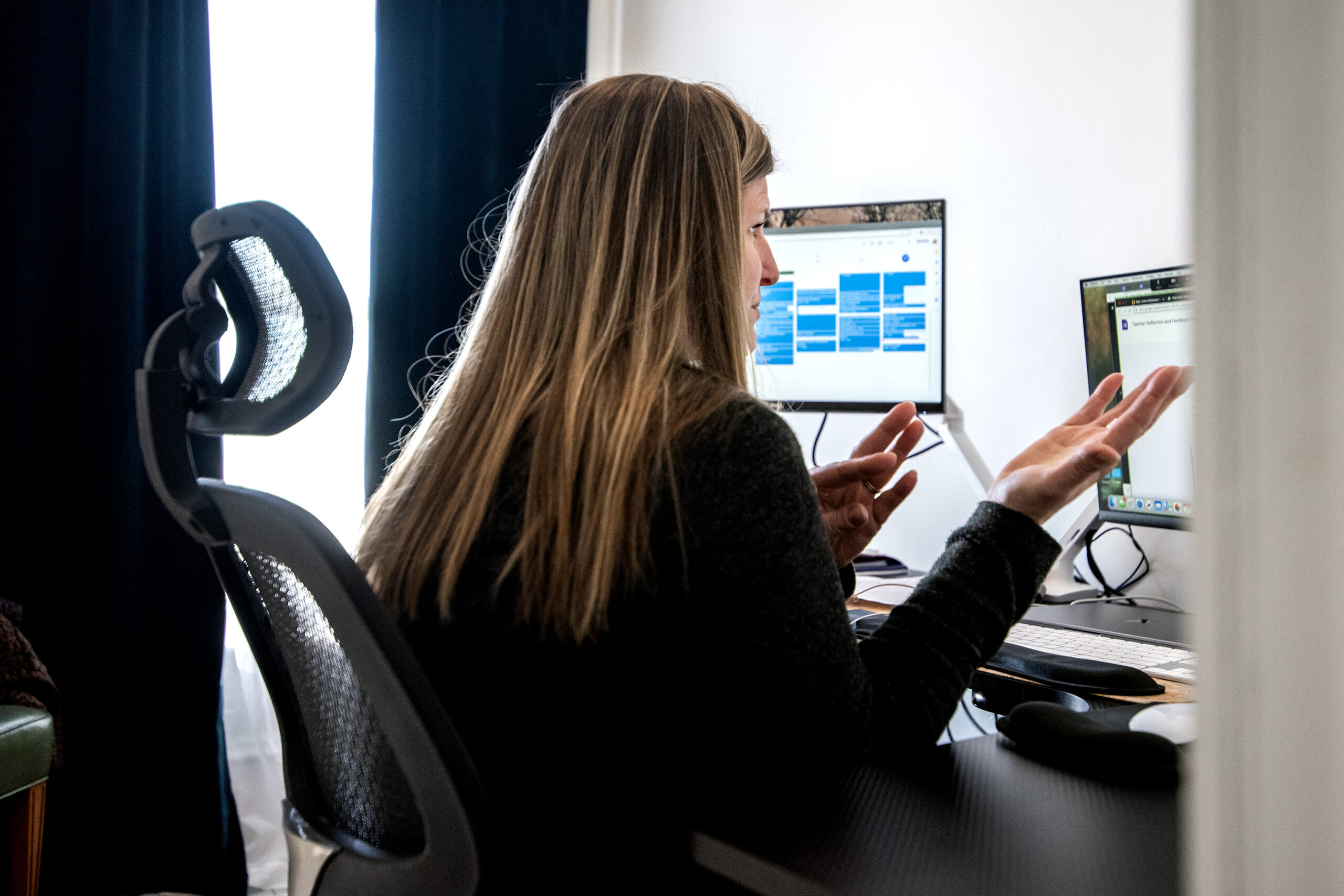 A woman is seen gesturing at a desk during a virtual meeting.