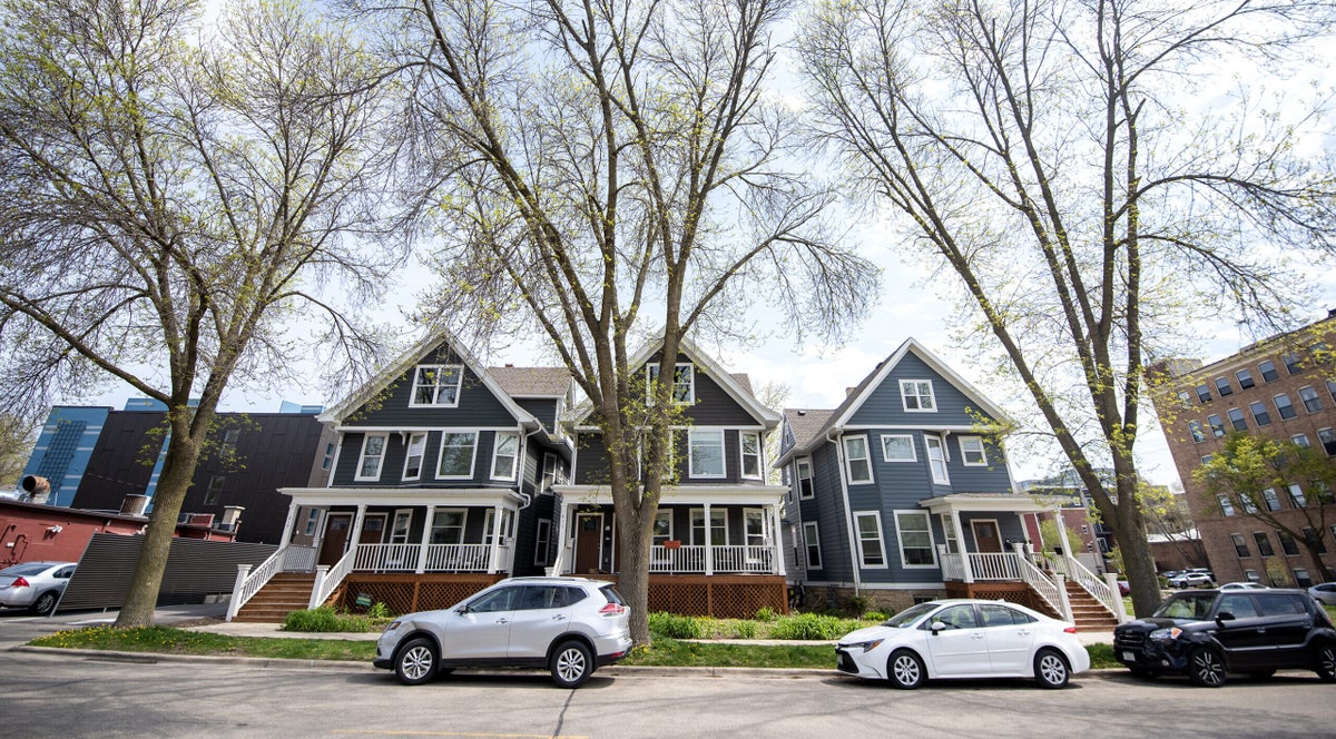 Tall trees are seen in front of multi-story homes. Cars are parked on the street in front of them.