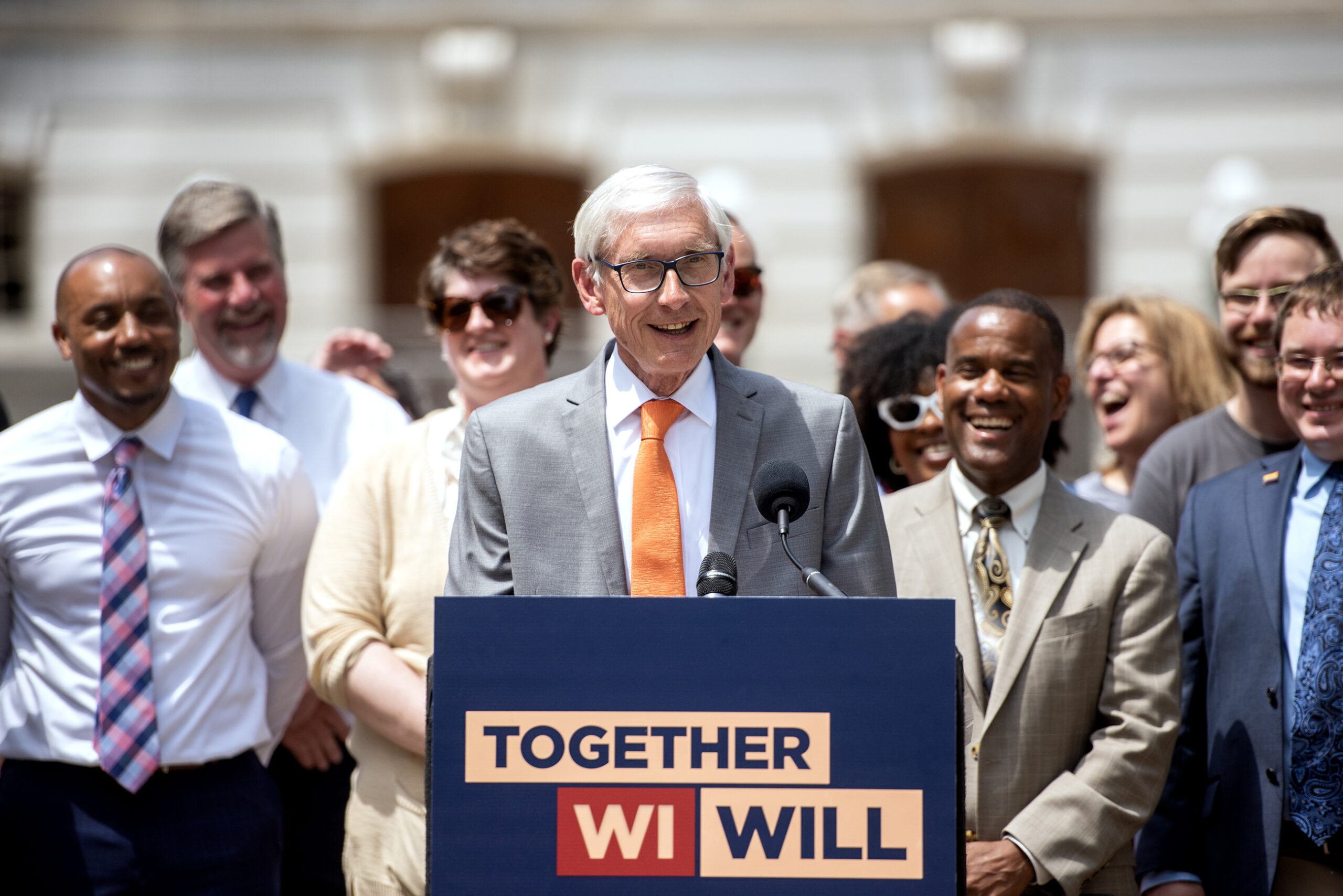 Gov. Tony Evers smiles as he speaks at a podium outside the Capitol building.