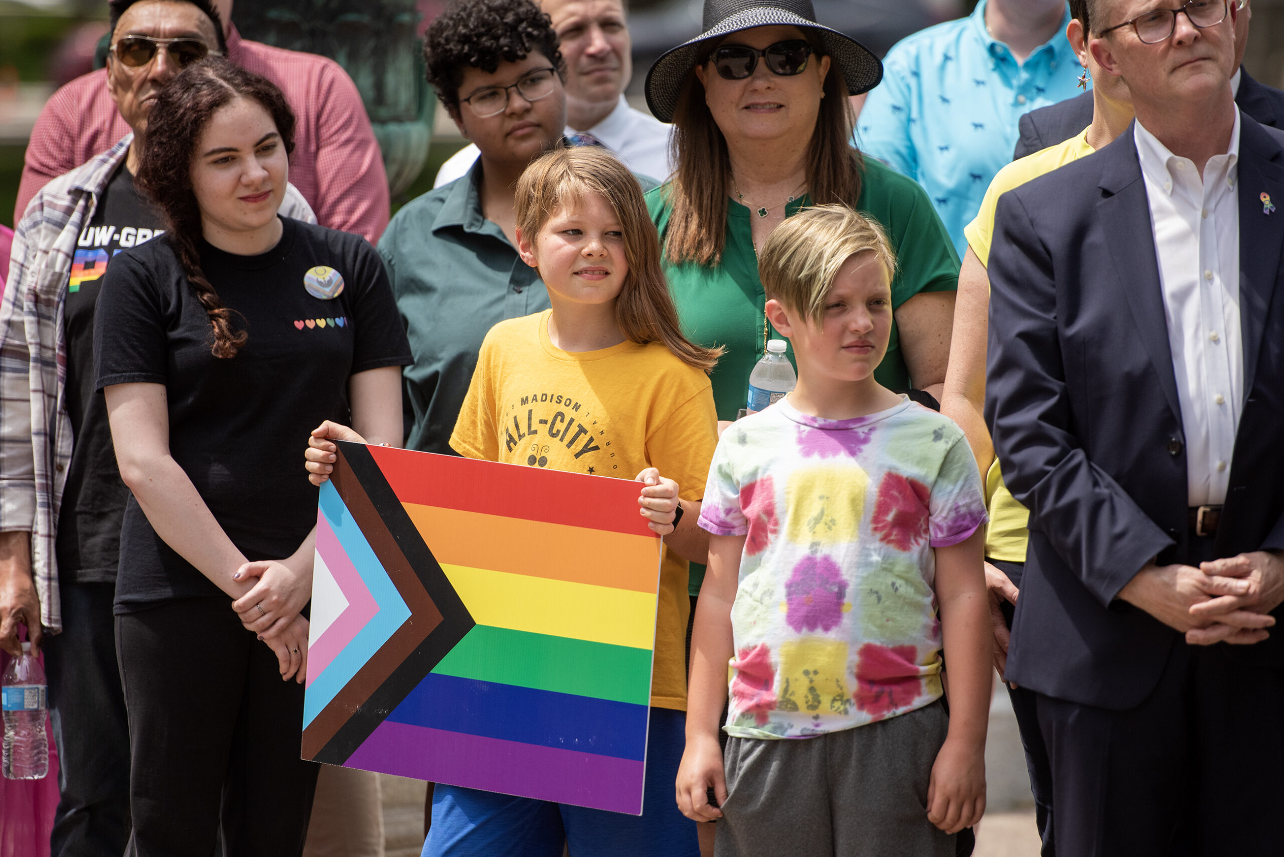 A group of people stand together outside the capitol. A child holds a Pride flag poster.