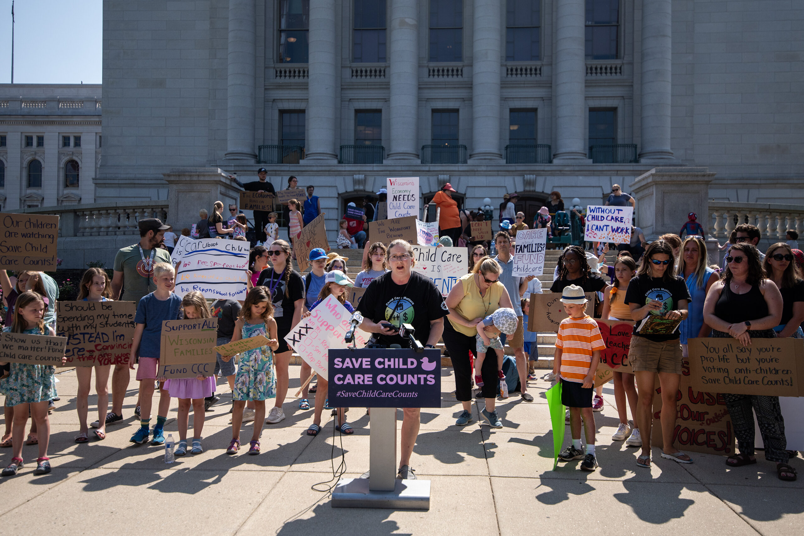A person stands at a podium with children, parents, and others behind her holding signs.