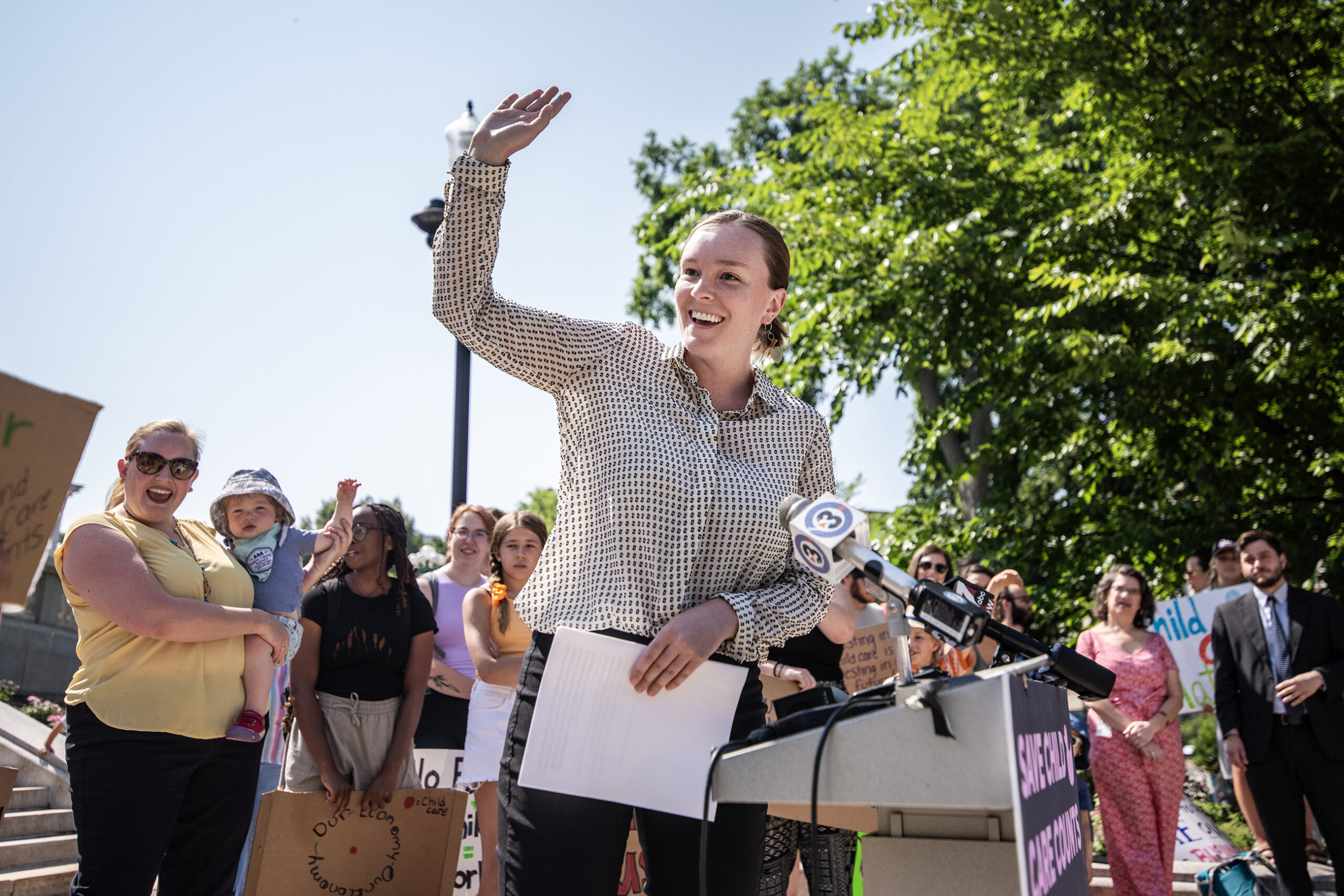 Rep. Greta Neubauer stands at a podium and gestures to the people behind her as she speaks outside.