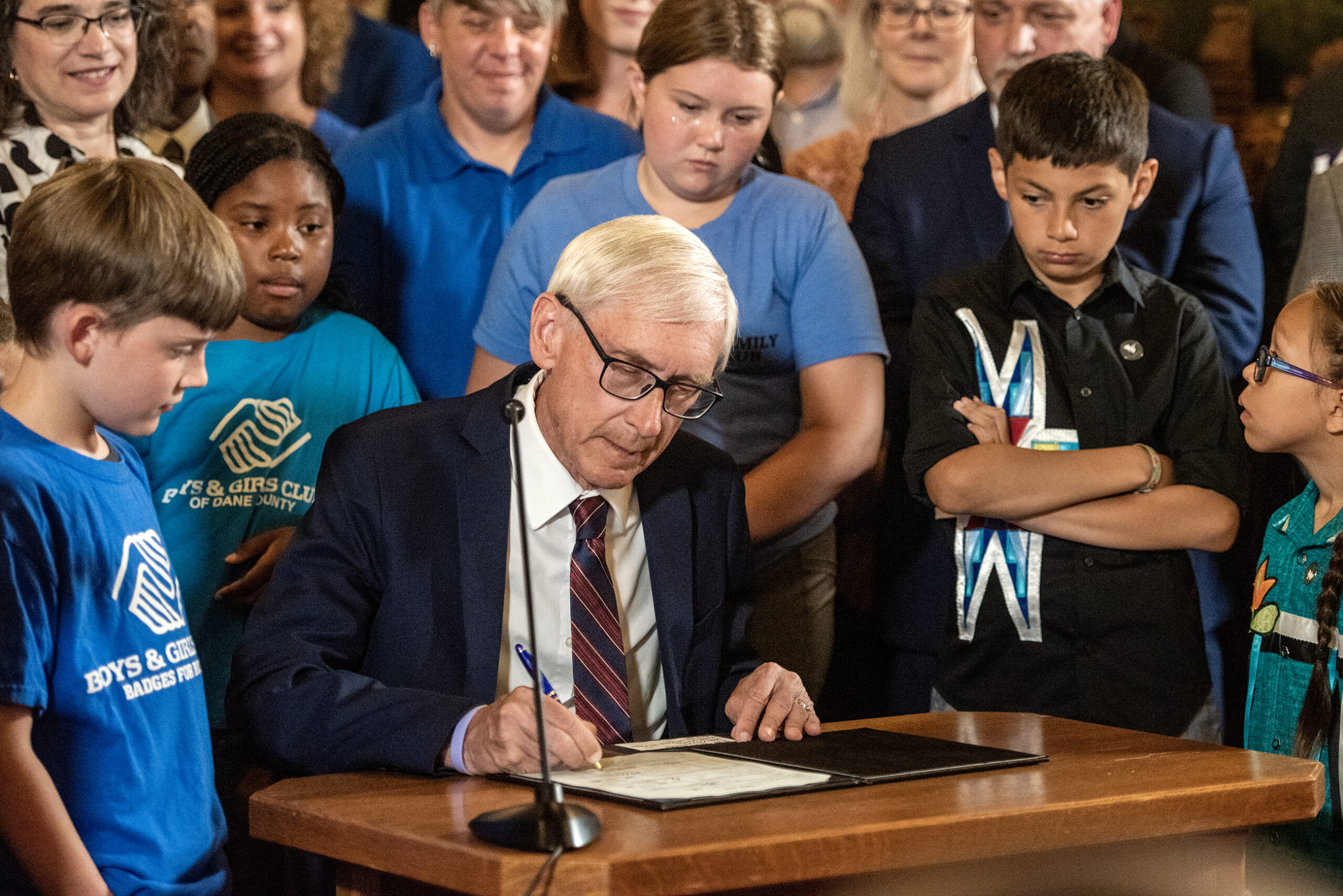 Gov. Tony Evers leans over a piece of paper as he signs it. Children attending the event watch over his shoulder.