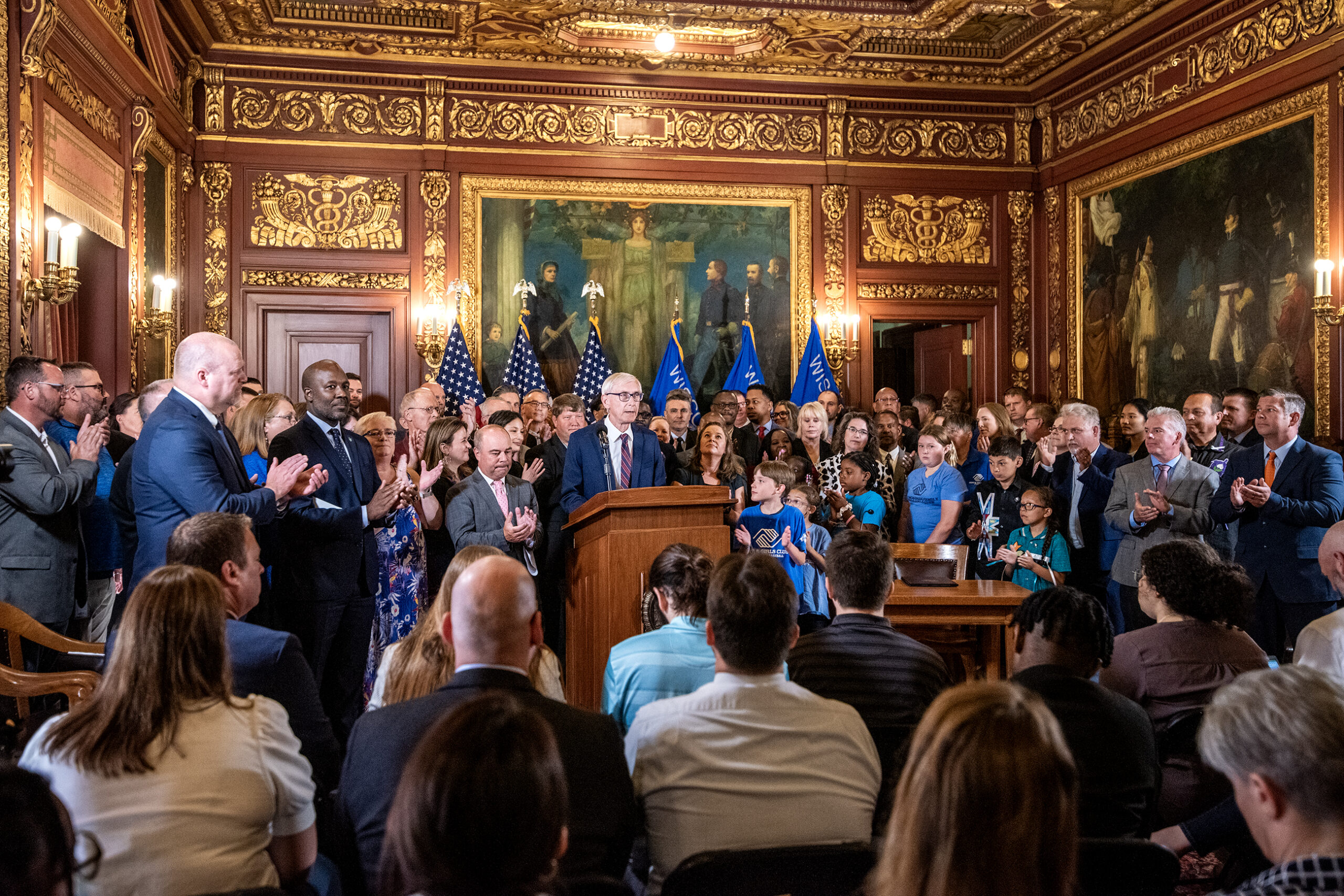A room in the Wisconsin State Capitol is filled with officials standing behind Gov. Evers speaking at a podium.
