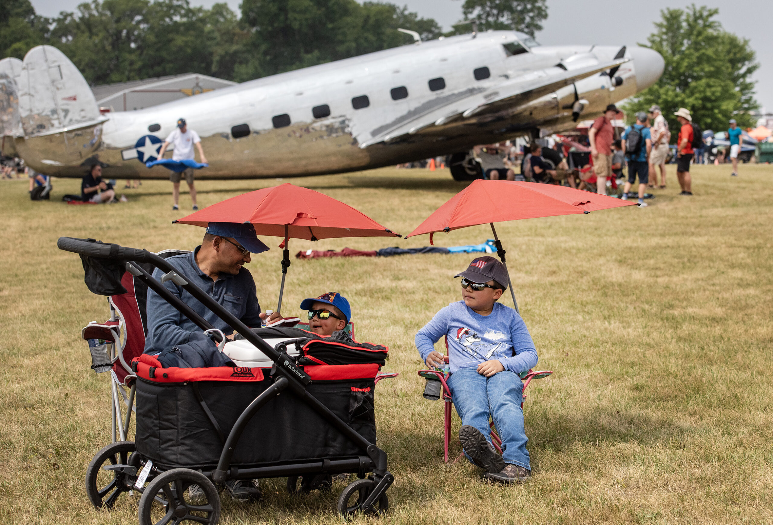 A dad sits with two small boys in lawn chairs underneath umbrellas. A large silver airplane can be seen behind them.
