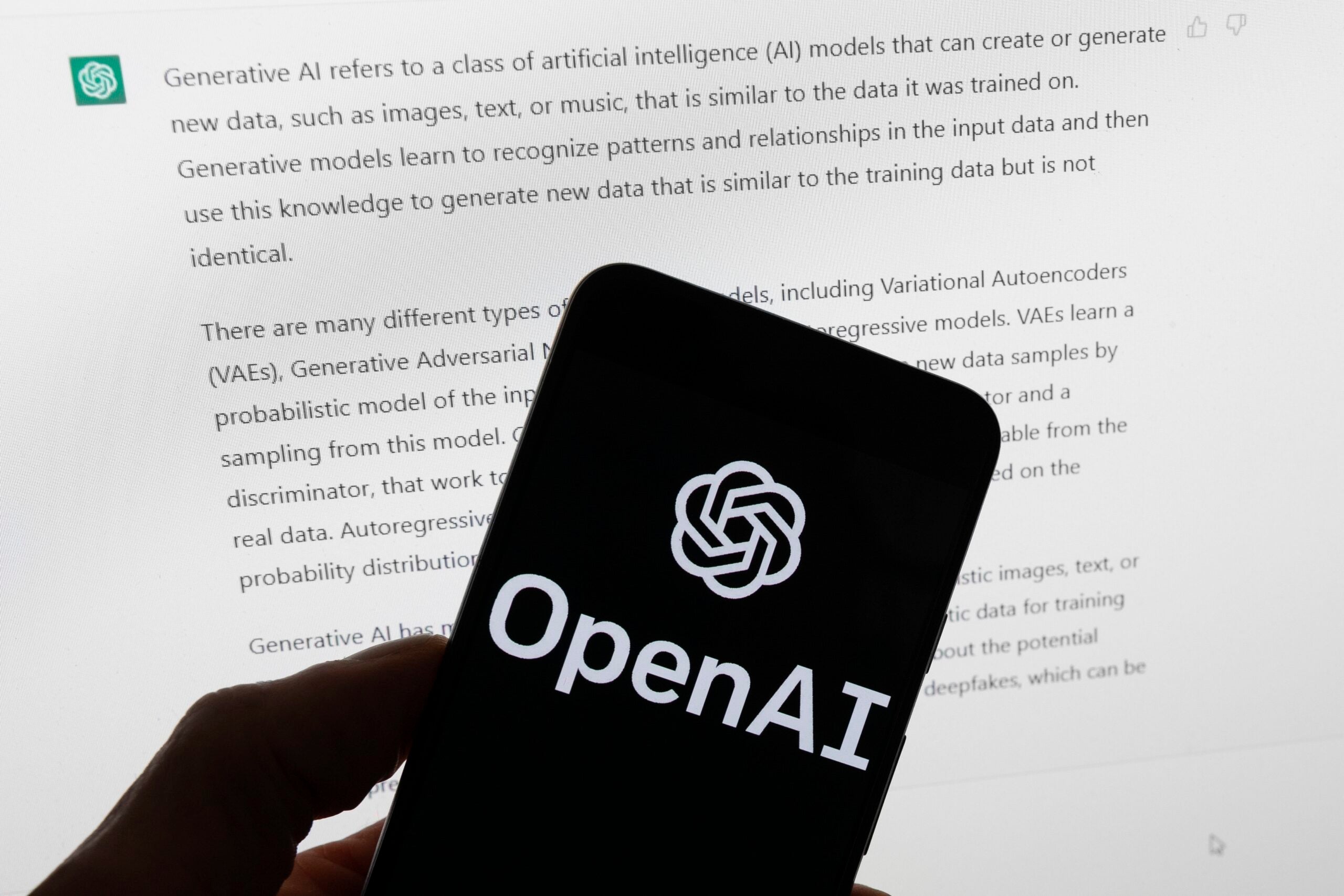 The OpenAI logo on a mobile phone in front of a computer displaying output from ChatGPT.