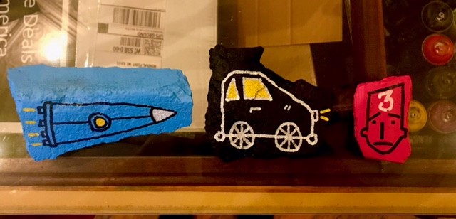 Left slab of concrete is blue with a spaceship, the middle slab is black with a white outline of a car, and the one on the right is pink with a face sketched out.