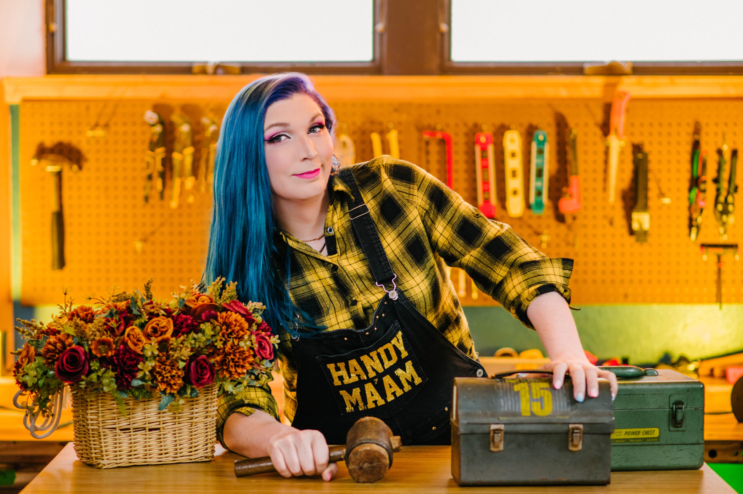 Mercury Stardust, a Trans woman with long blue and purple hair, wears overalls that say "Trans Handy Ma'am" in gold glitter lettering. She leans on her elbow with her hand resting on an old-fashioned toolbox and poses smiling at the camera.