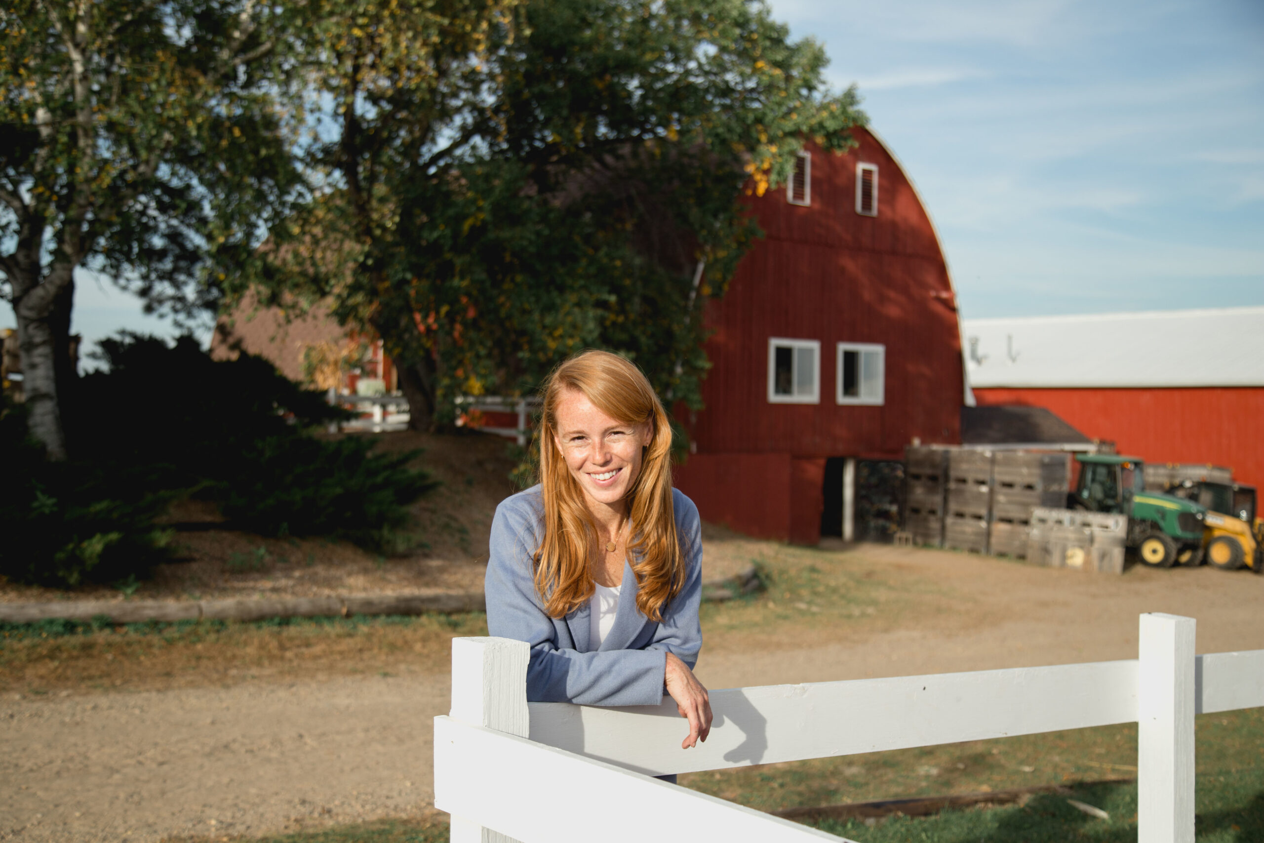 Rebecca Cooke leans against a white fence in front of a red barn