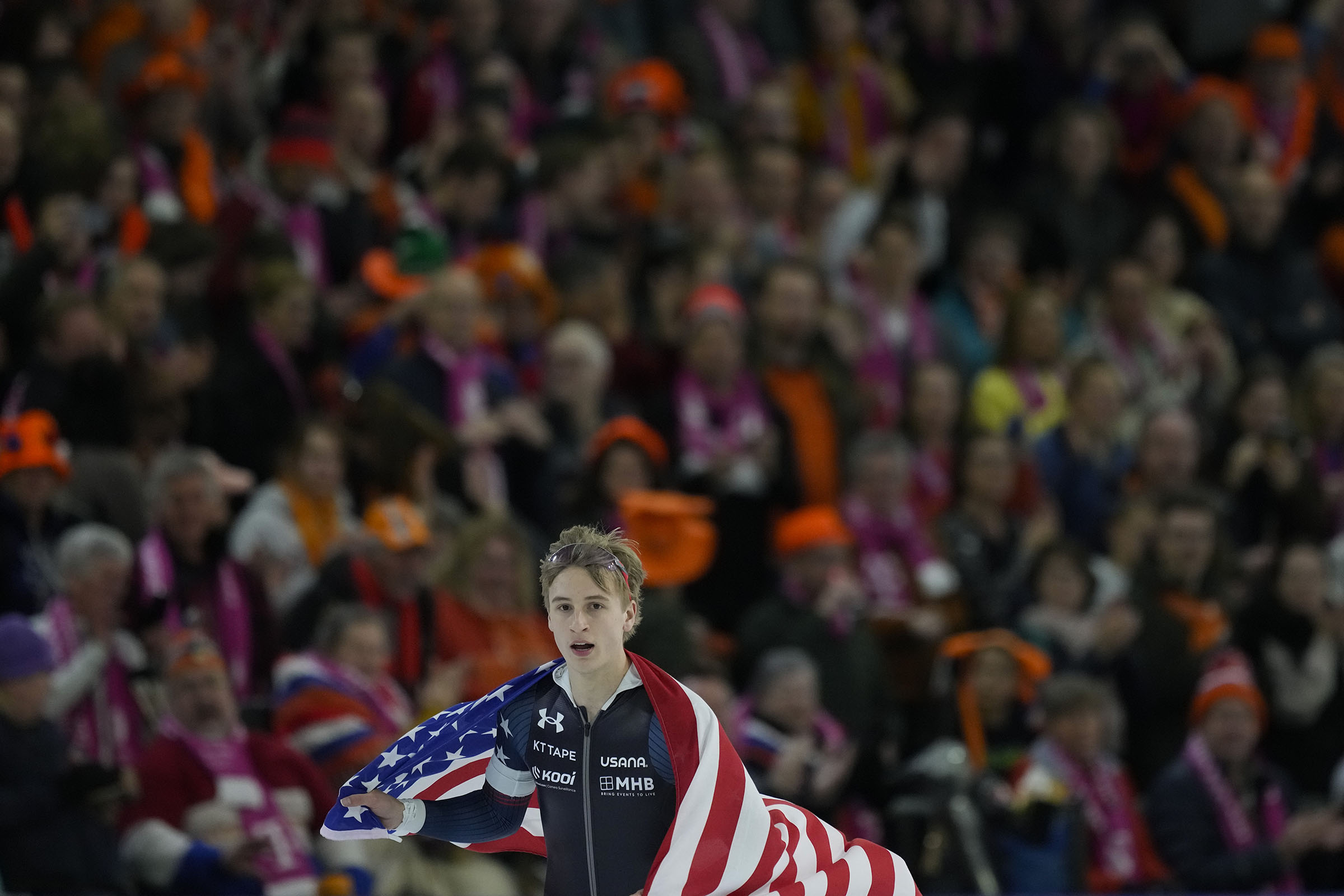 A speedskater holds an American flag in front of a crowd in a Dutch arena following a gold-medal win