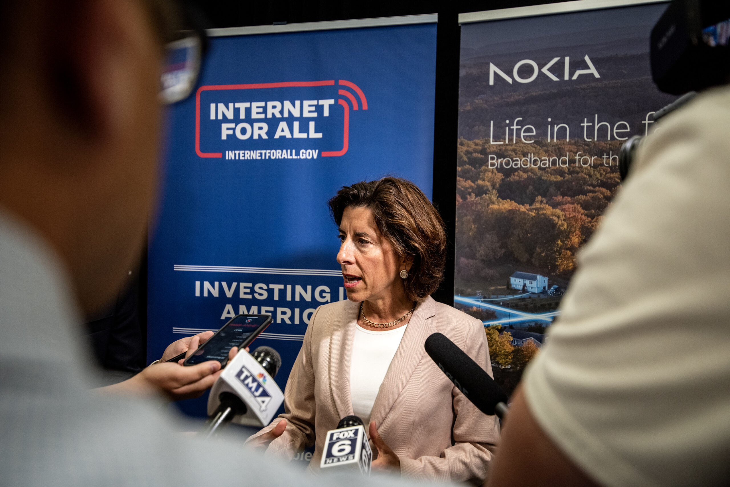 Sec. Of Commerce Gina Raimondo stands as reporters hold microphones to hear her speak.