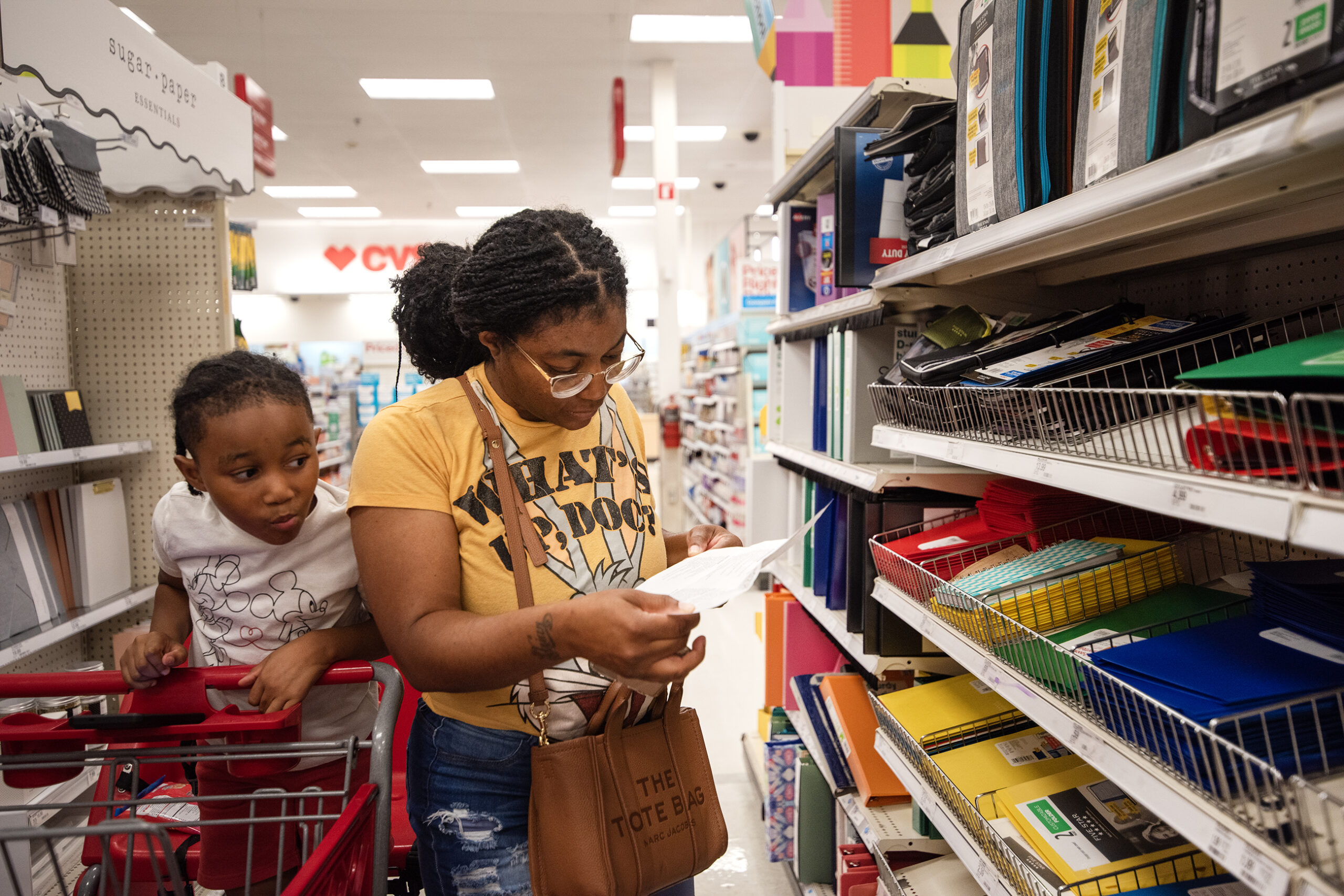 A shopper looks at a paper list while her son looks over her shoulder from the shopping cart.