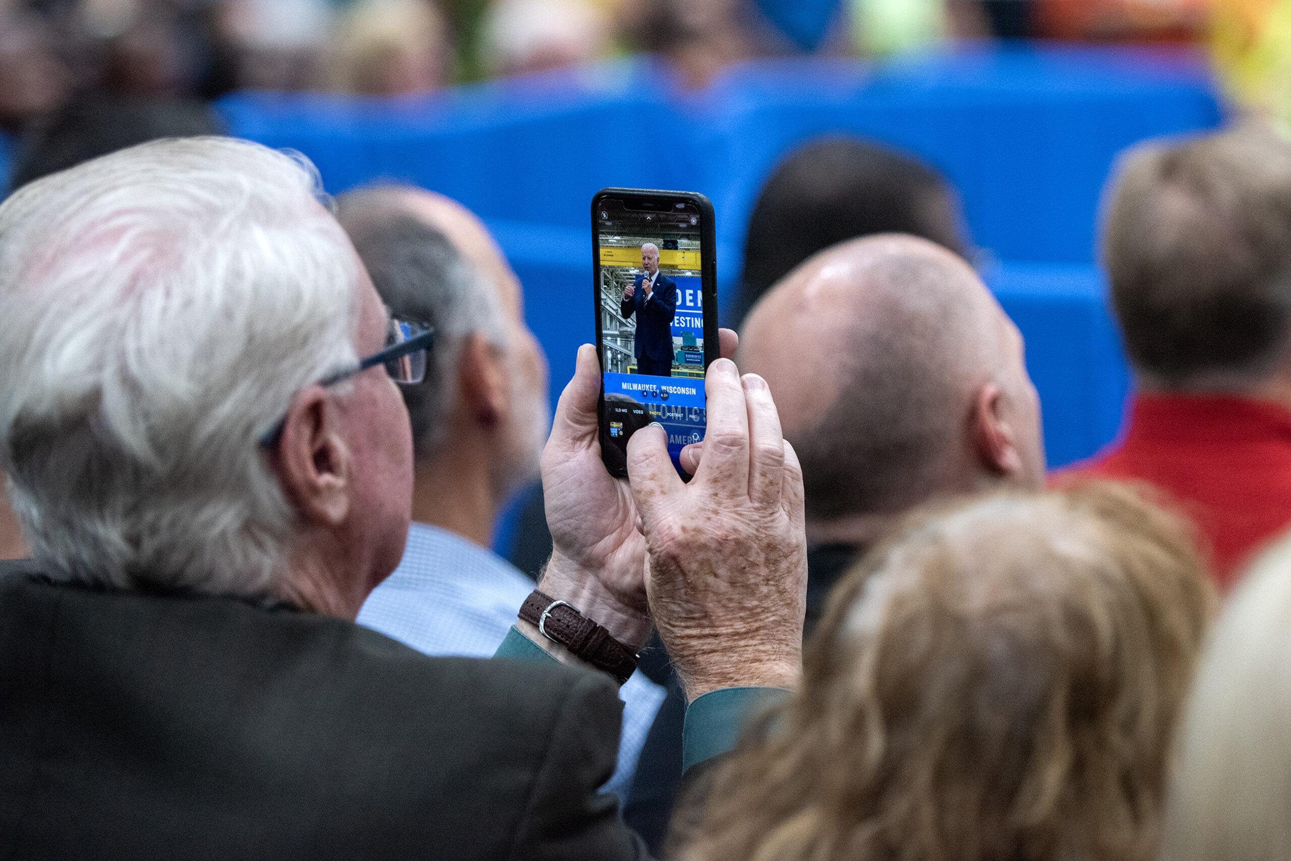A man holds a phone while he takes a photo of President Biden, who can be seen on his screen.