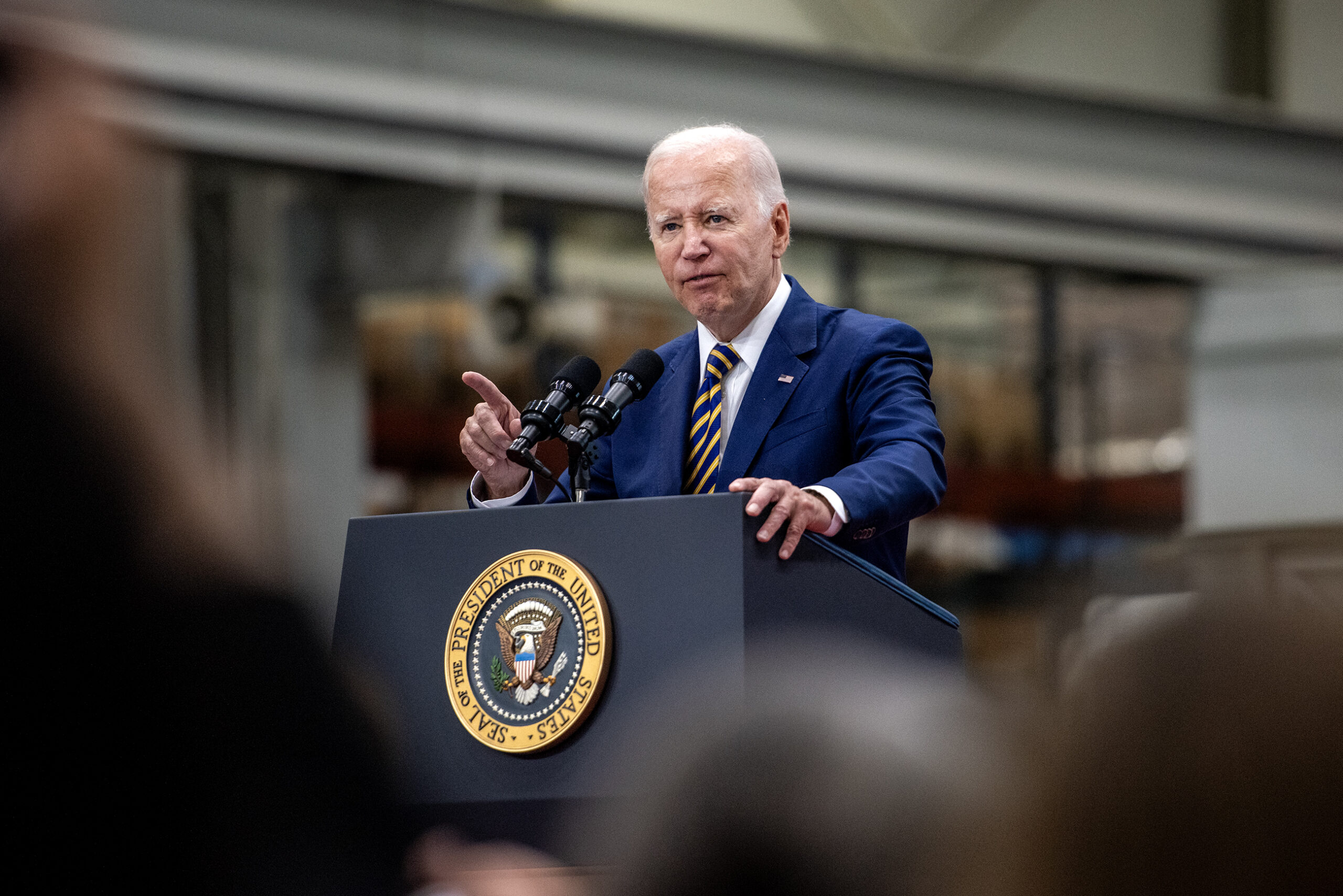 President Biden can be seen above the heads of attendees as he speaks at a podium.