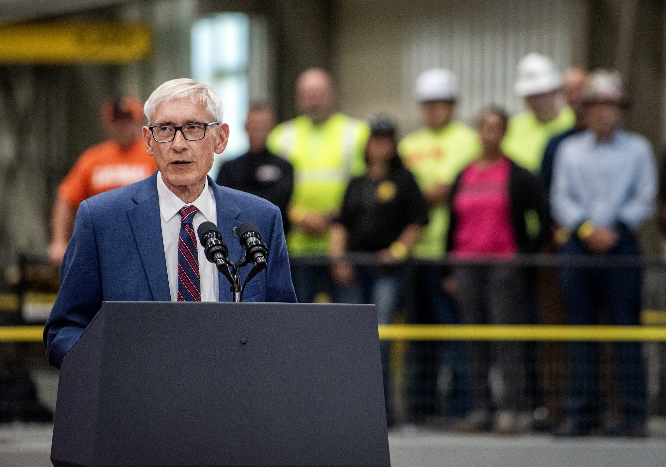 Gov. Tony Evers speaks at a podium. Workers can be seen behind him.