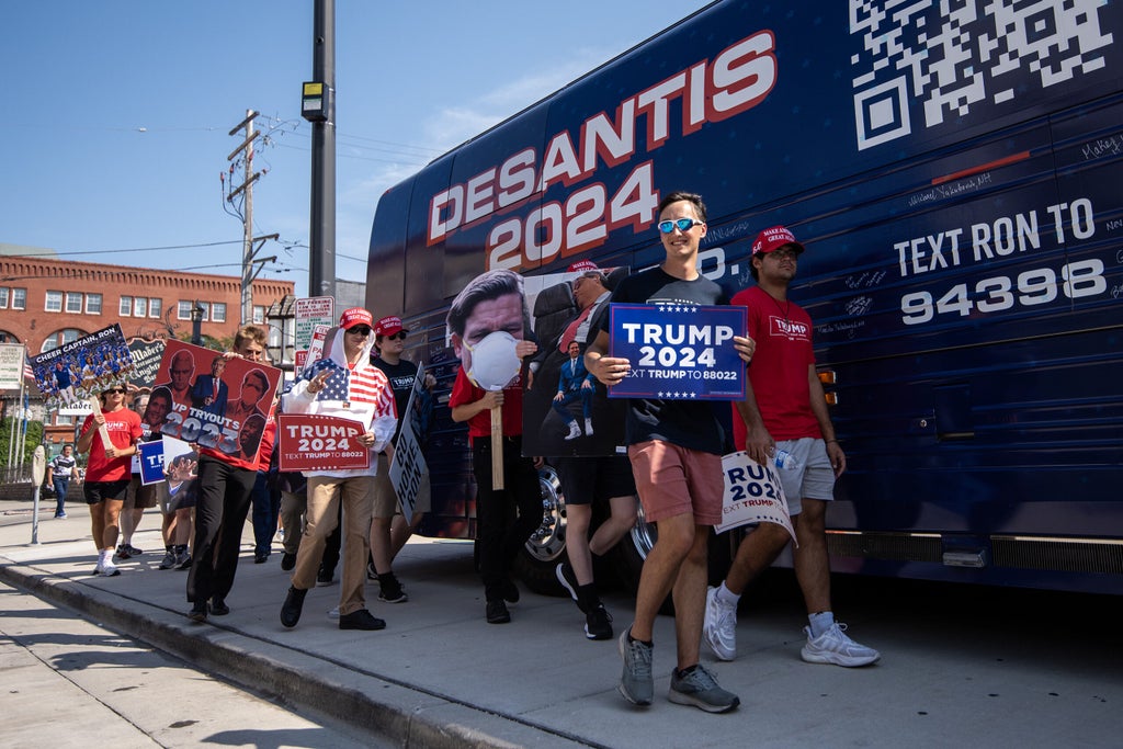 A blue van with the words "DeSantis 2024" is parked as protesters with Trump signs walk by.