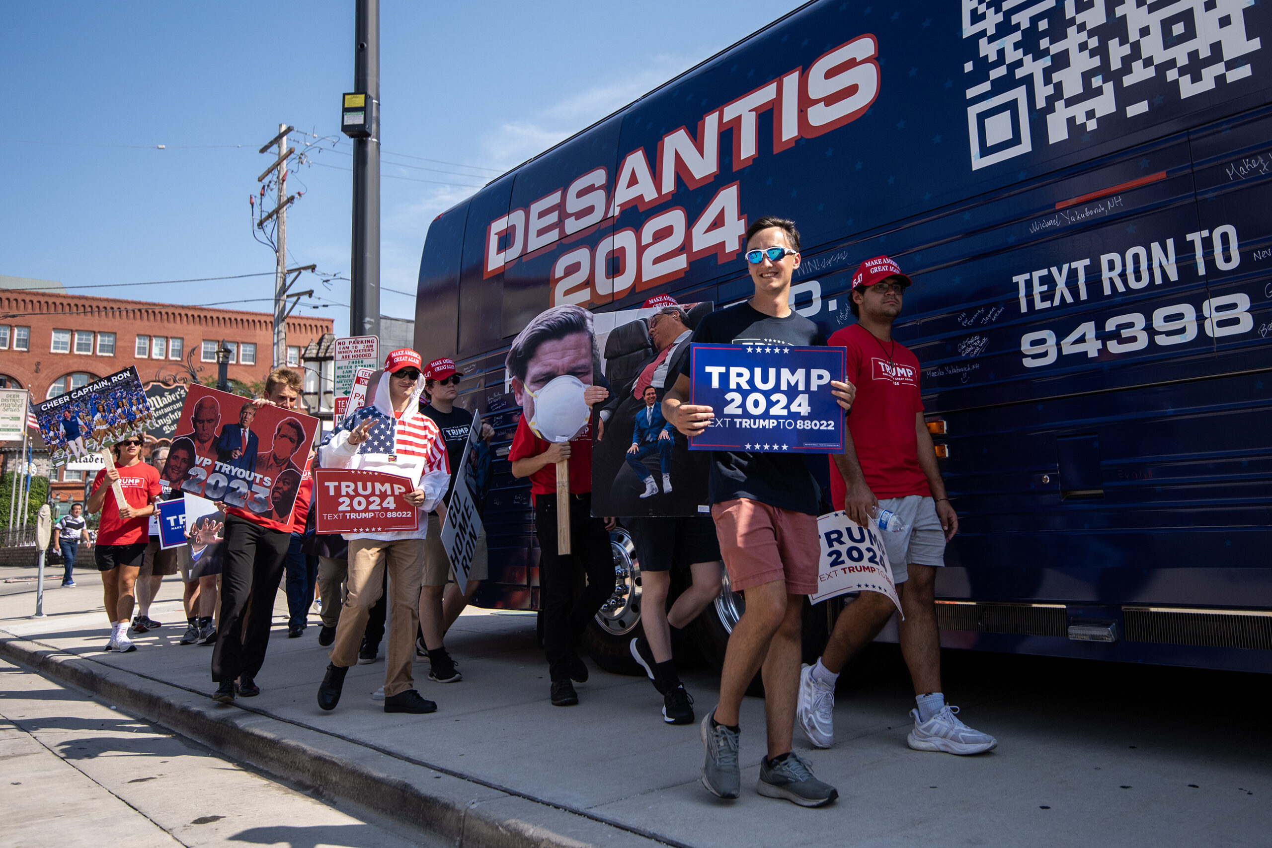 A blue van with the words "DeSantis 2024" is parked as protesters with Trump signs walk by.