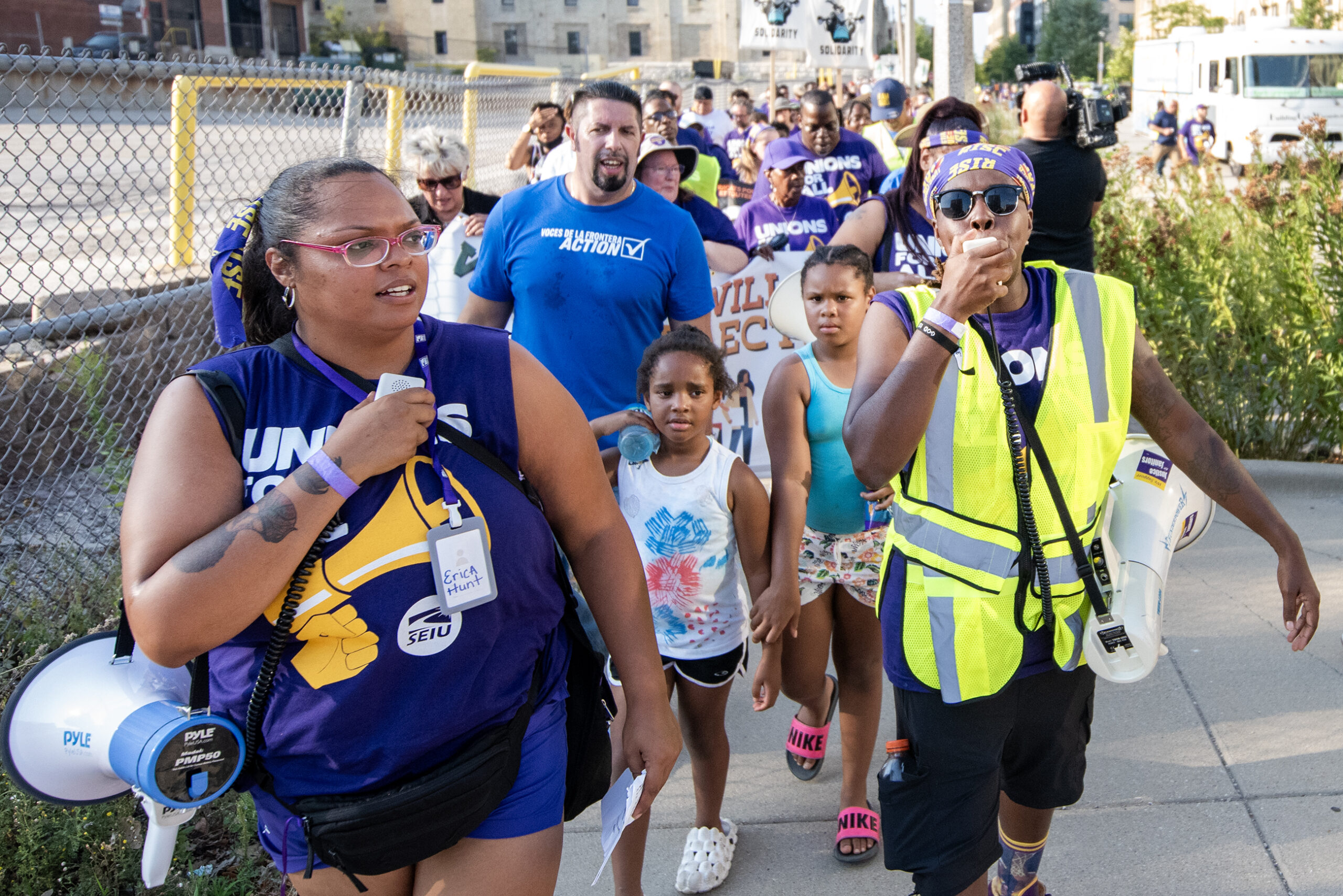 Protesters in matching purple shirts march on the sidewalk behind a leader in a neon vest.