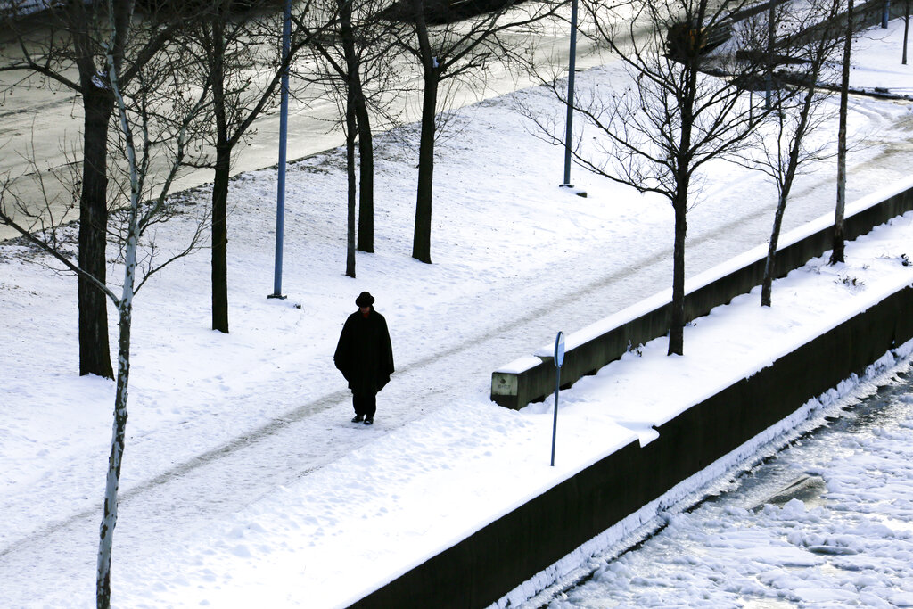 A person dressed in winter clothes walks down a snow-covered sidewalk next to a frozen river.