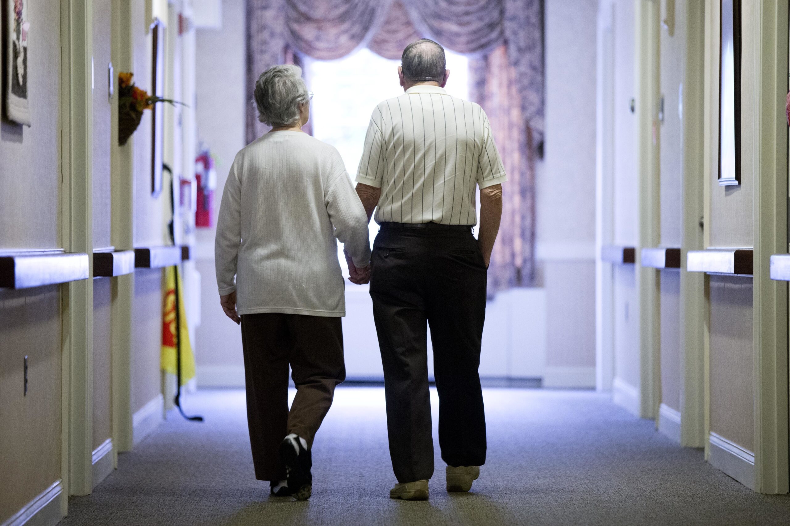 An elderly couple walks down a hall of a nursing home