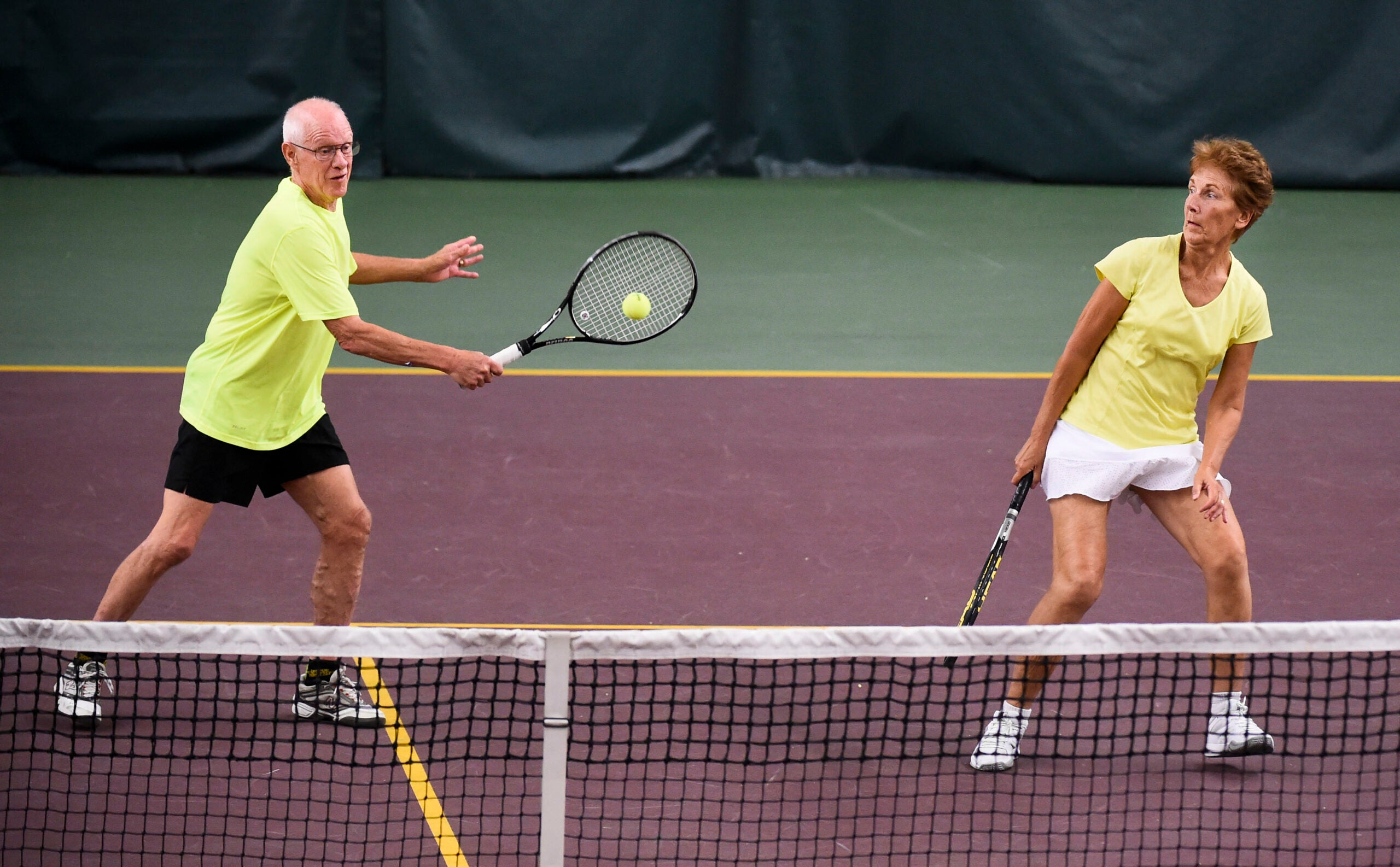 An elderly man hits a tennis ball with his raquet while his partner, an elderly woman, stands next him.