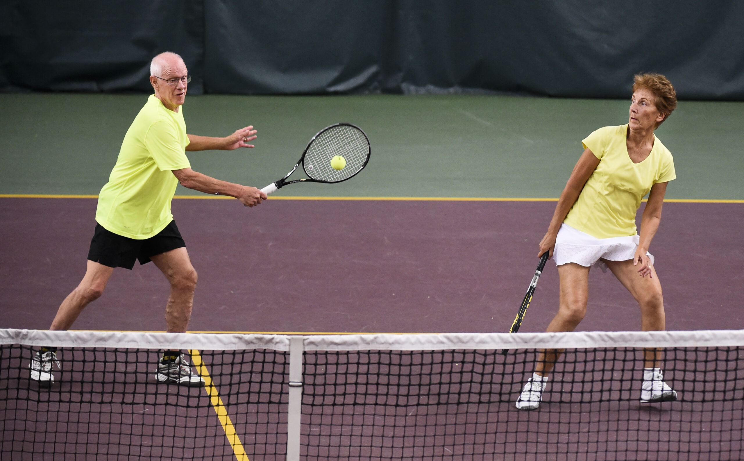 An elderly man hits a tennis ball with his raquet while his partner, an elderly woman, stands next him.