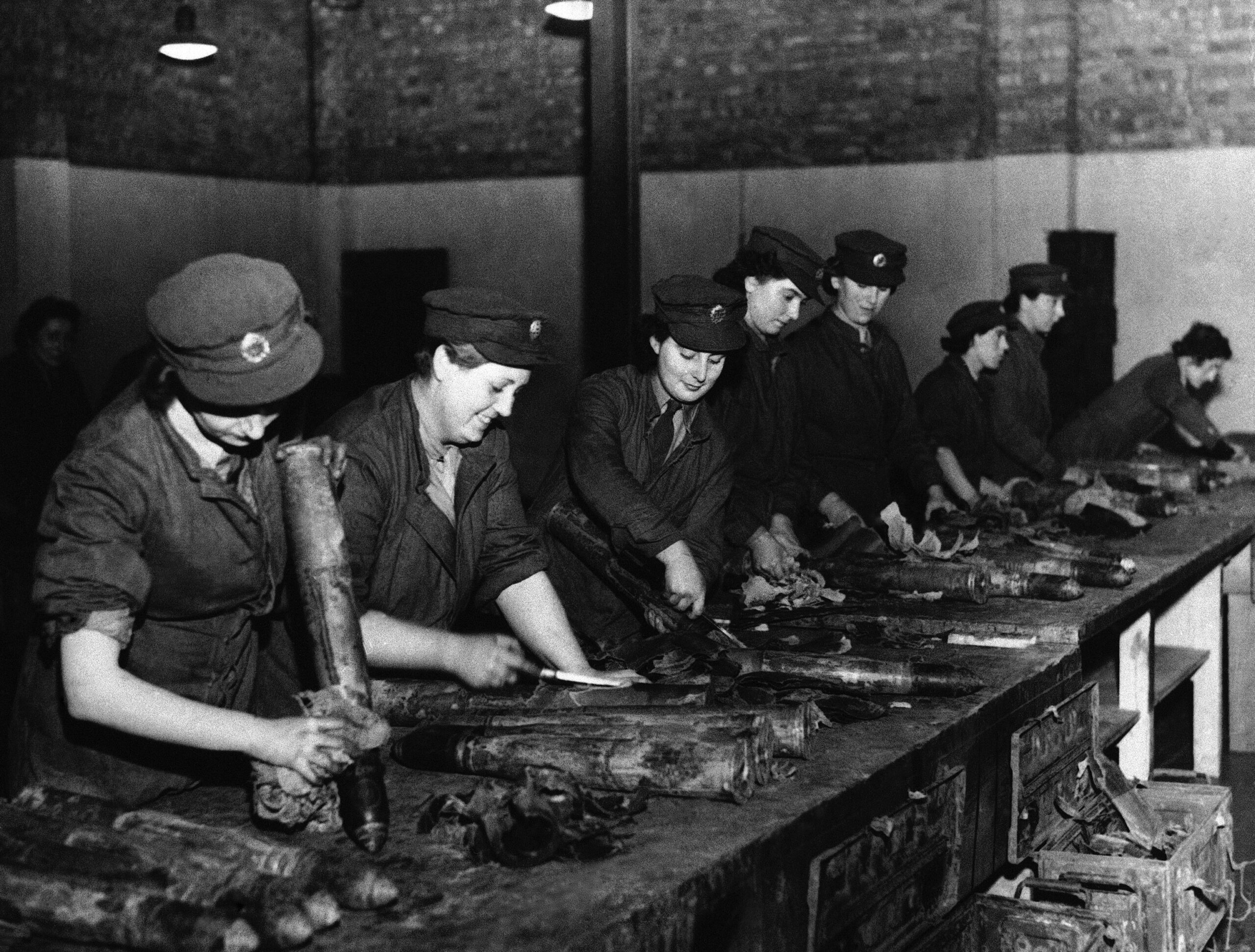 In this old photo, women in factory uniforms handle dirty bomb shells. Some of them wipe them clean with a rag.
