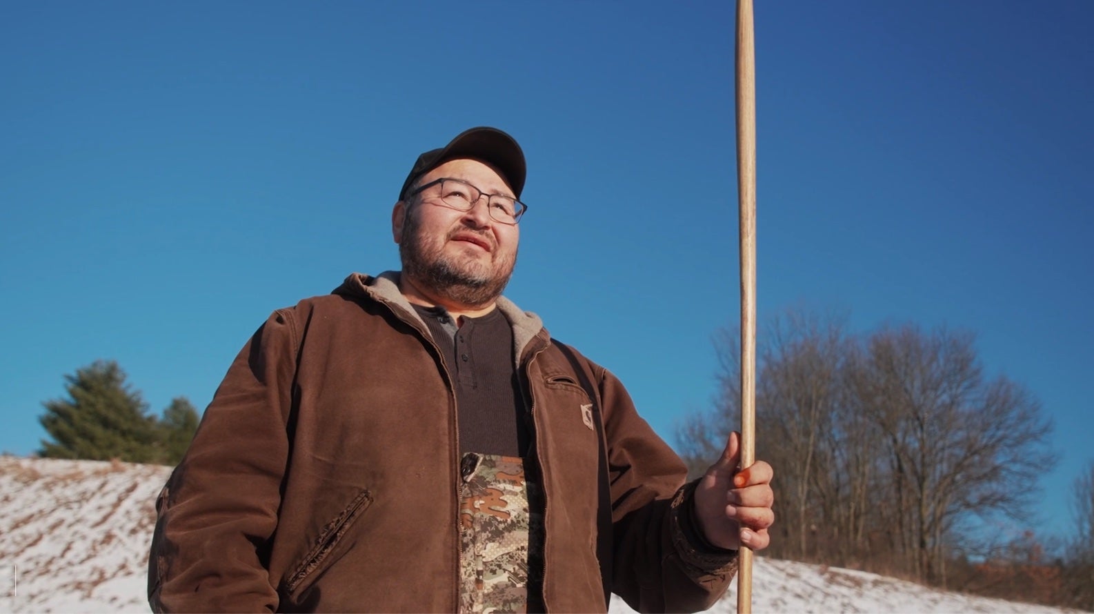 A man in a brown jacket stands holding a wooden rod upright. Behind him is a snowy hill and leafless trees in Winter.