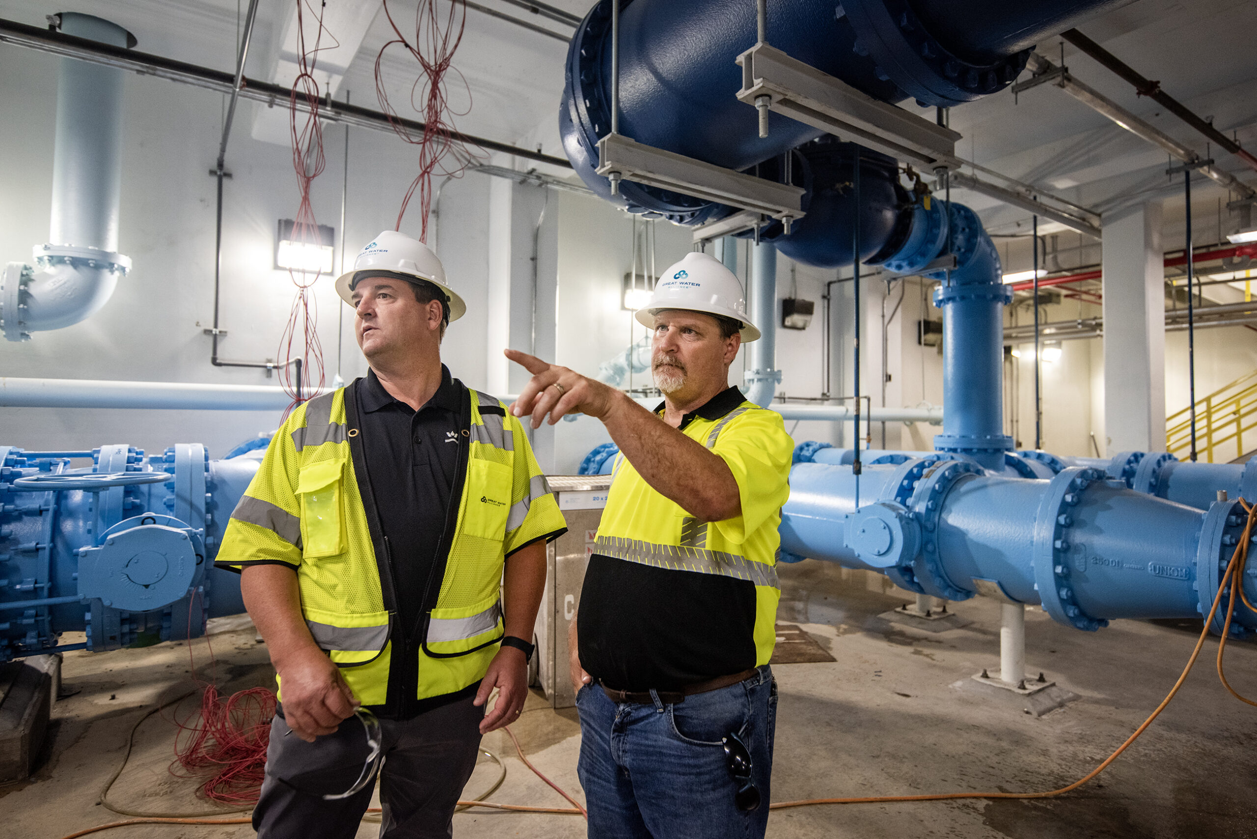 Two men in hard hats and neon vests stand in a room filled with pipes.