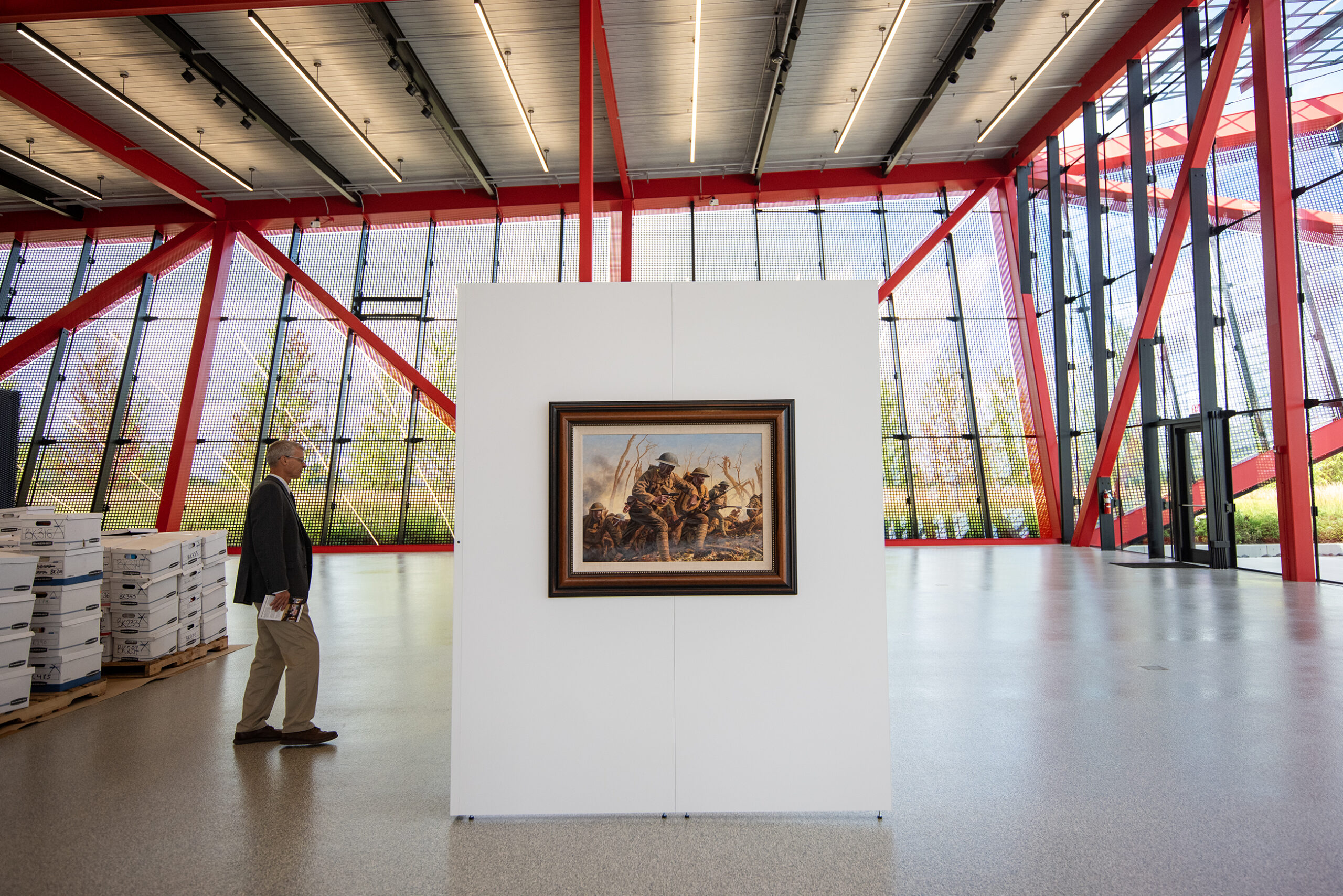 A framed piece hangs on a small white wall inside a large space. The building's red beams can be seen in the background.