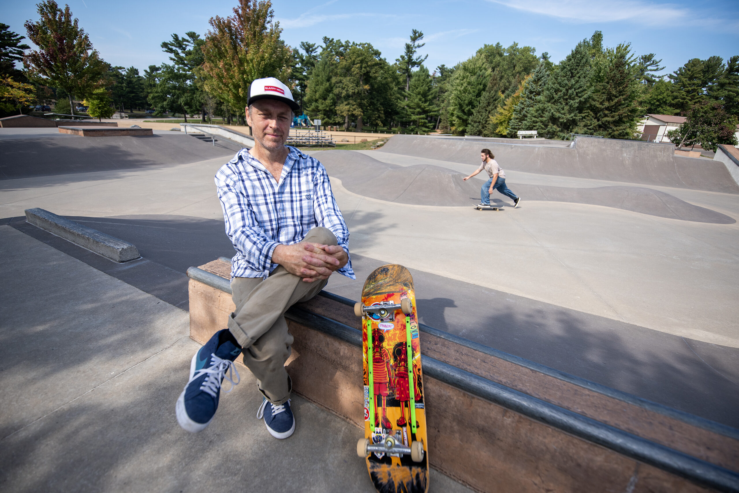 John sits on the side of a skate park next to his skateboard. A man skateboards behind him.