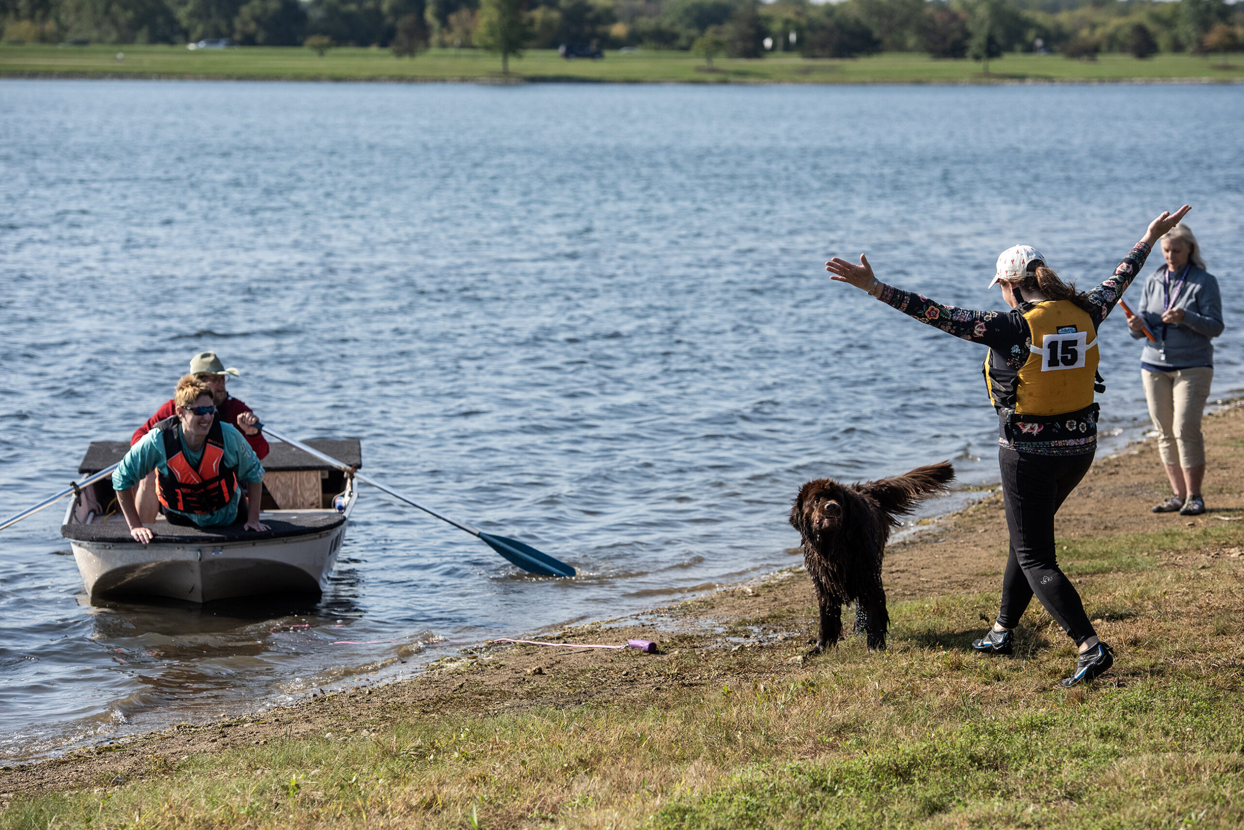 A dog stands on the shore of a lake as a woman approaches with her arms raised excitedly.