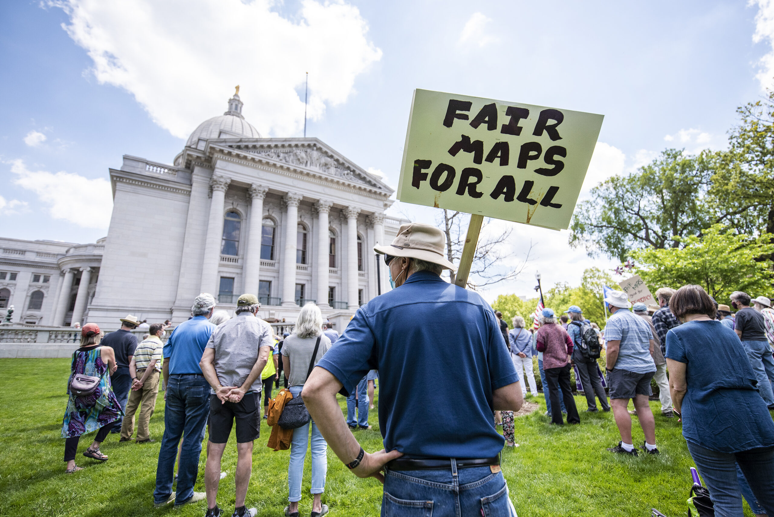 People stand outside the Wisconsin State Capitol on a sunny day. A man holds a sign that says "FAIR MAPS FOR ALL."