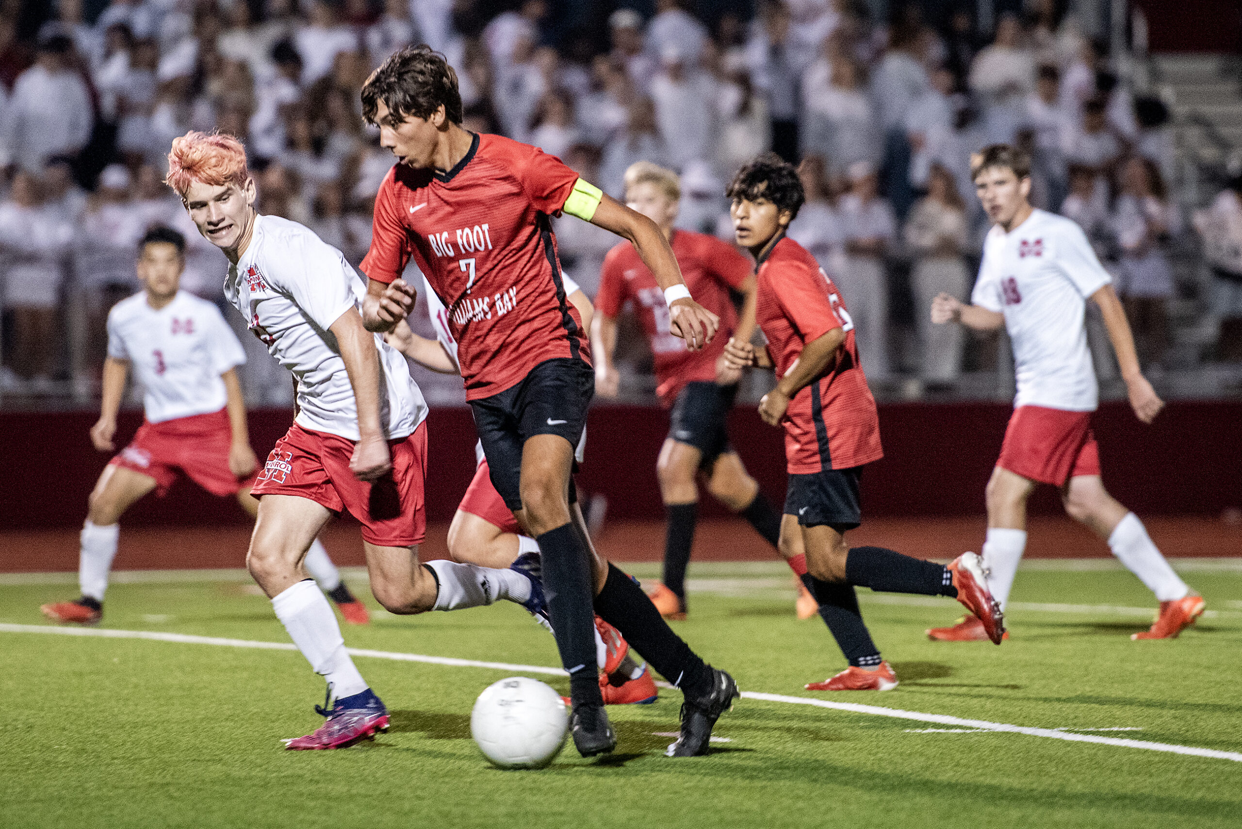 A soccer player in a red jersey kicks the ball as opposing players approach.