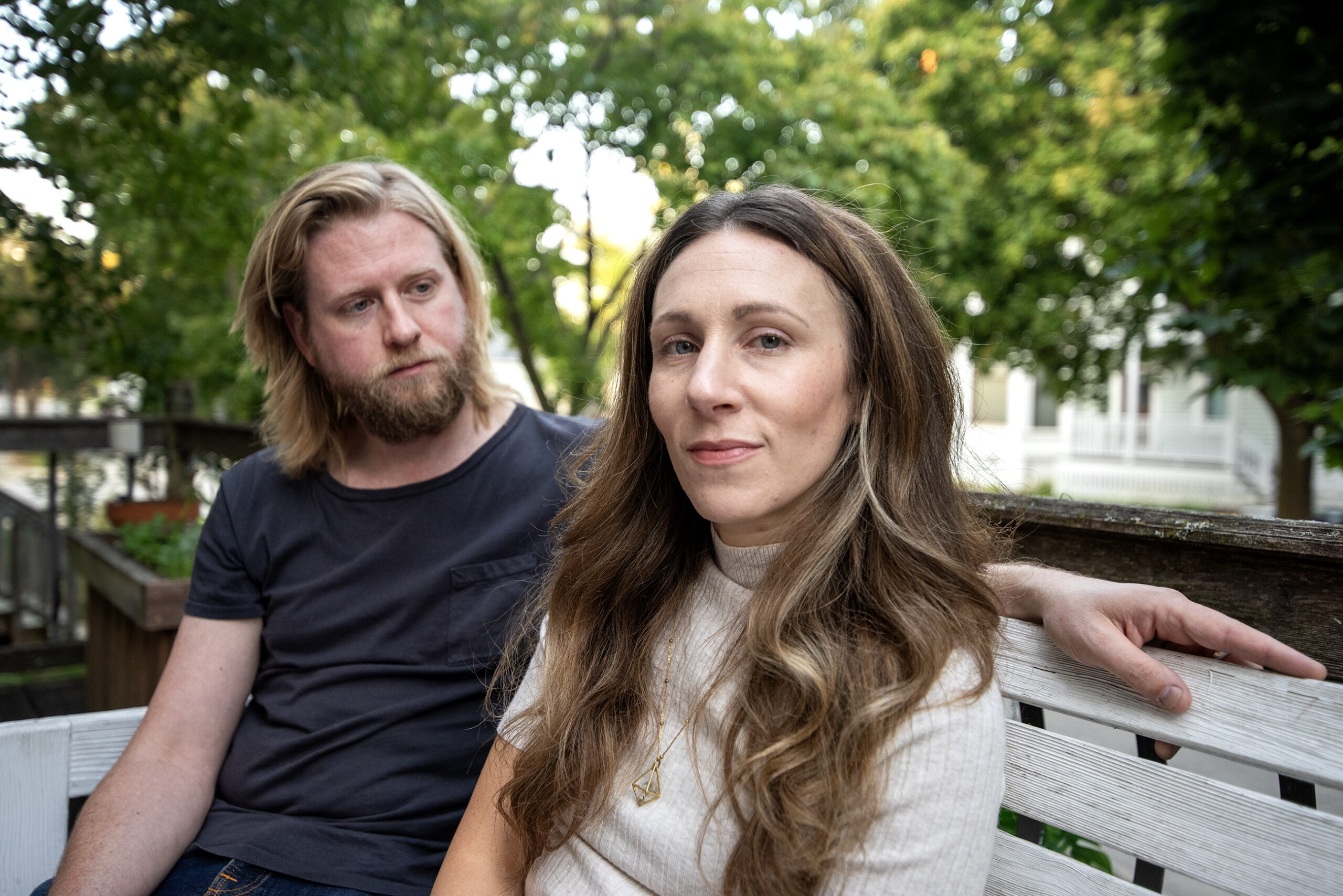 Steve looks at Megan as they sit together on a bench. Green leaves on trees can be seen behind them.